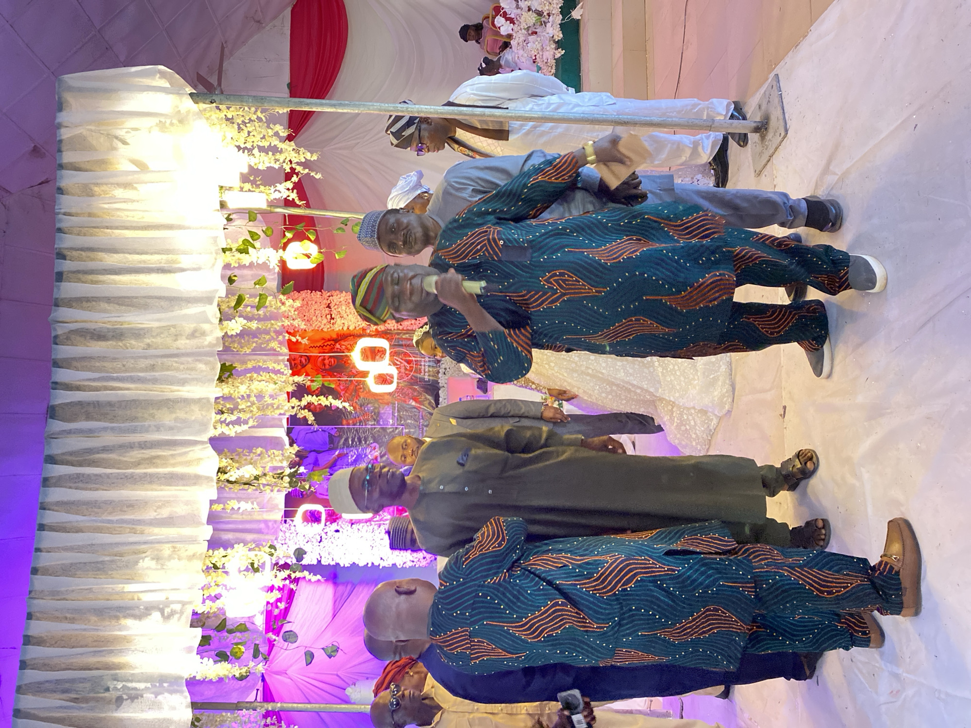 The image shows a group of men standing in what appears to be a decorated indoor space, likely in Dutsen Kato, Nigeria. The men are dressed in traditional Nigerian attire, with some wearing hats and flowing gowns. One man is holding a microphone, suggesting he may be speaking or performing. The backdrop is decorated with white and red fabric, floral arrangements, and some light fixtures. The lighting in the space is a mix of natural and artificial, casting shadows. The floor is a light color, possibly tile or marble.