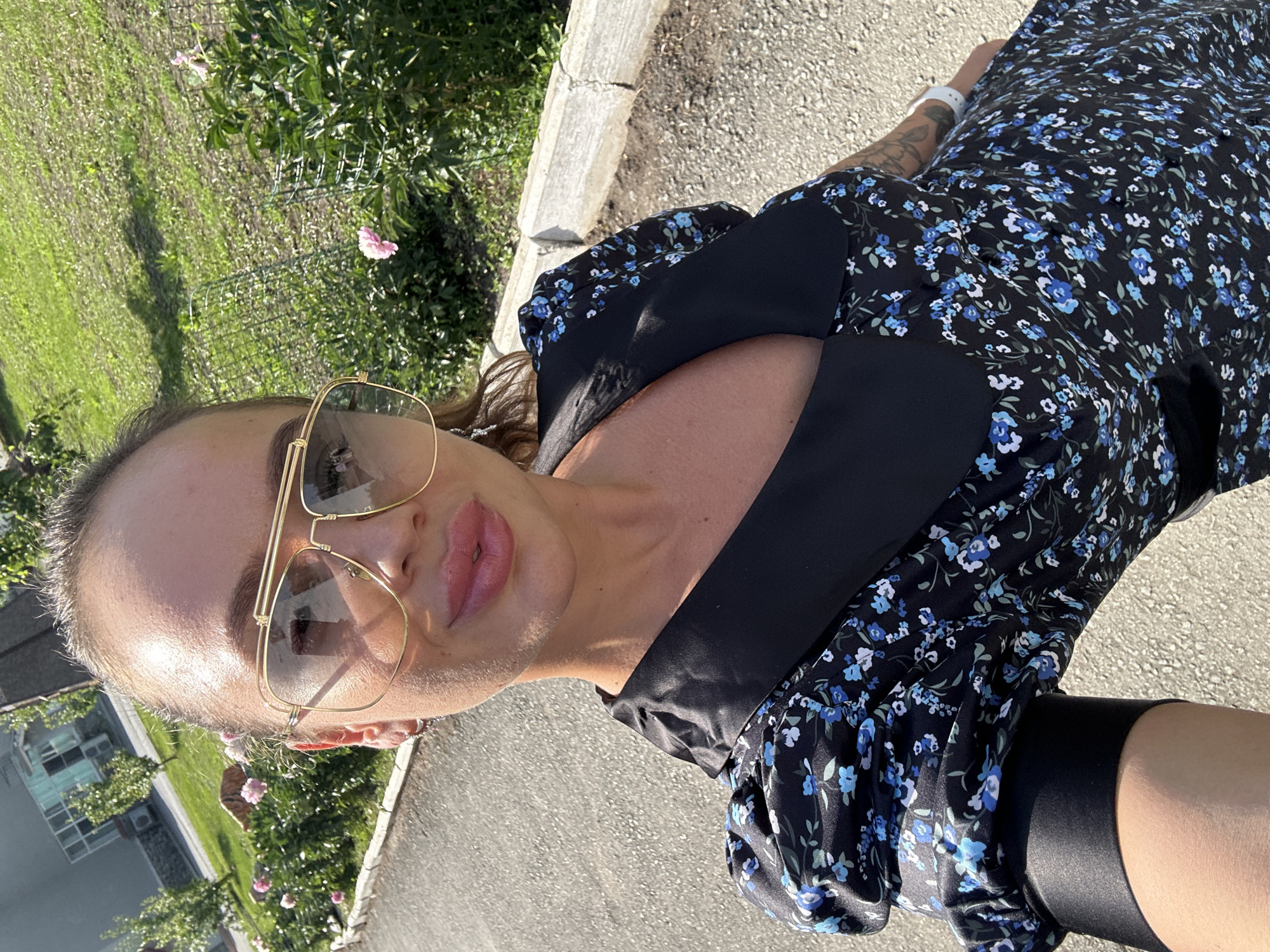 The image shows a woman taking a selfie outdoors. She is wearing a black floral dress with blue flowers and large gold-rimmed sunglasses. The setting appears to be a sunny day, with greenery and a paved surface visible. The woman's pose and the close-up nature of the selfie suggest a casual and personal moment. The lighting indicates it is daytime, possibly late afternoon due to the warm tone of the sunlight. The background includes some vegetation and what looks like a building in the distance. The woman has a tattoo on her arm, and she is wearing a white wristband. The overall mood is relaxed and perhaps happy. No text is visible in the image.