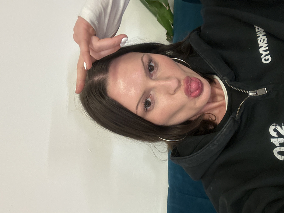 The image shows a person, likely a woman, captured in a playful and casual pose. Her head is tilted significantly, and she is looking directly at the camera with her tongue sticking out slightly. She has medium-long dark brown hair, parted down the middle, and fair skin. Her eyes are open, and she appears to be wearing some mascara. Large silver hoop earrings are visible in both ears.
One hand, with fingernails painted white, is positioned above her head, with the index and middle fingers crossed, a common gesture for good luck or an "X" shape.
She is wearing a dark-colored, likely black, zippered hoodie. A white collar or neckline of an undershirt is visible beneath the hoodie. On the upper right side of the hoodie, the brand name "GYMSHARK" is clearly visible in white capital letters. Further down on the right side of the hoodie, partial white numbering, possibly "012" or similar, is also present.
The background is simple, featuring a plain white wall or surface on the left side of the frame. On the right, there is a dark teal or blue object, possibly a cushion or part of a couch. A small portion of a green plant is barely visible in the upper right corner. The overall impression is one of lightheartedness and an informal moment. The image itself is oriented sideways, with the subject's head towards the top of the frame.