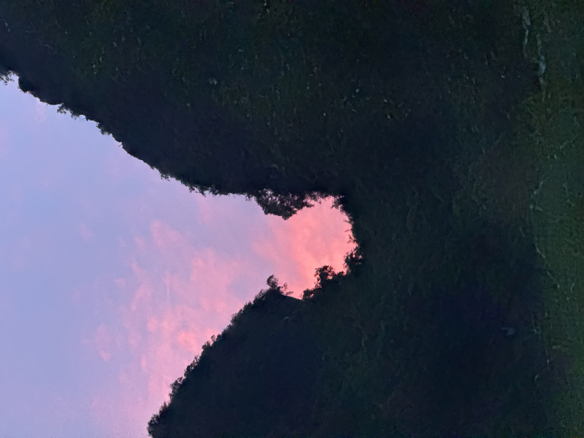 A serene evening view captures a gap between two silhouetted mountains, revealing a vibrant pink and purple sunset sky. The mountains are covered in lush greenery, appearing dark against the colorful sky. The gap between them forms an irregular, organic shape, framing the celestial display. The overall scene is one of natural beauty and tranquility, suggesting a remote, picturesque landscape. The image is taken from a low angle, looking upwards towards the sky.