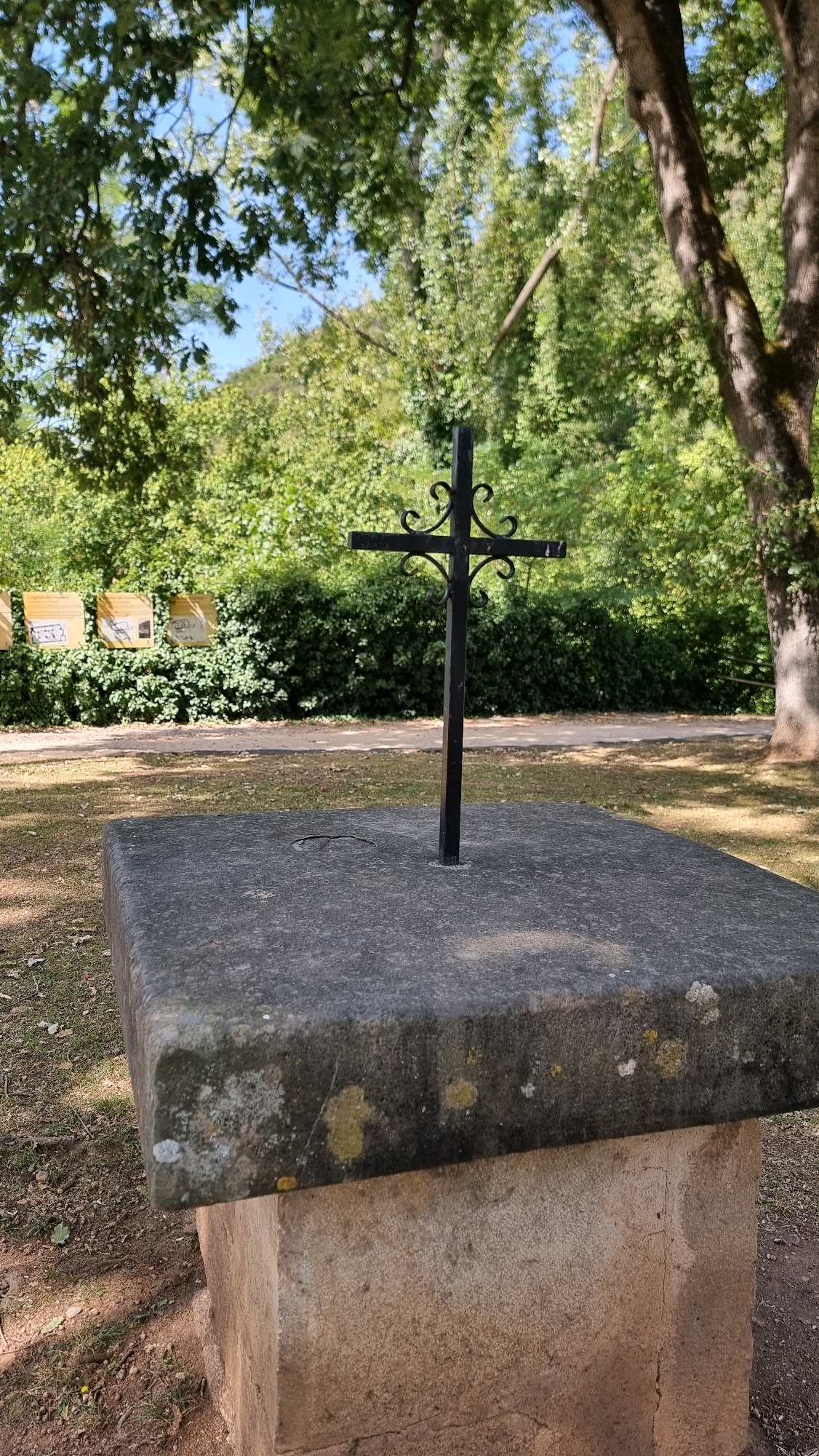 A black metal cross with decorative scrollwork stands upright on a large, flat stone pedestal in an outdoor setting. The pedestal appears weathered and is set on a lighter-colored base. Behind the pedestal and cross, lush green foliage and trees form a dense backdrop, with patches of blue sky visible through the leaves. In the mid-ground, a paved path runs horizontally, and to the left of the path, several yellow signs with text are mounted on a low hedge. The ground in the foreground and around the pedestal is covered in dry leaves and sparse grass. The overall scene suggests a peaceful, possibly historical or memorial site, likely in a park or natural area within the municipality of Sant Joan les Fonts, Spain.