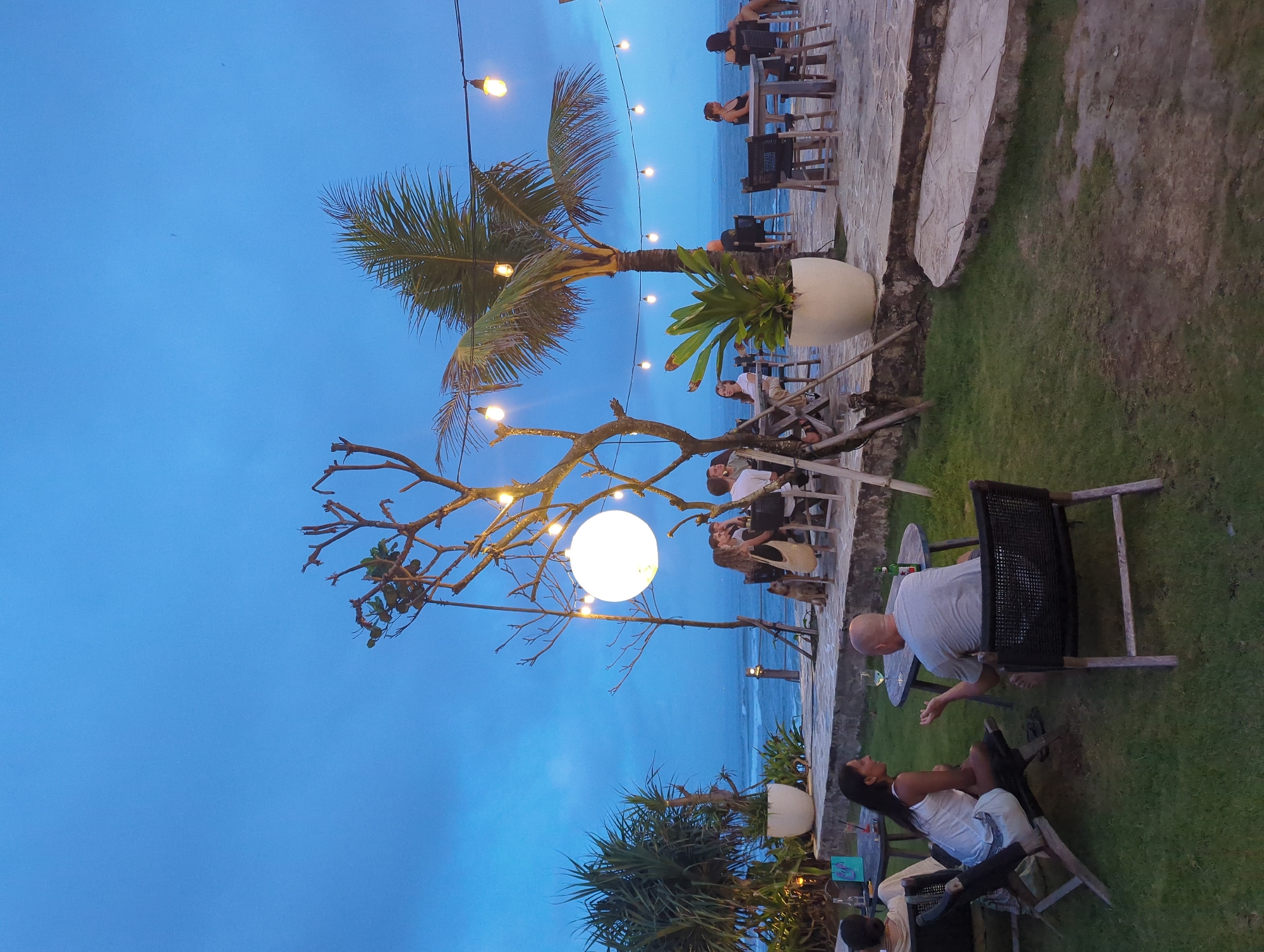 The photograph shows an outdoor dining area at dusk, likely in Mengwi, Indonesia. Several people are seated at tables and chairs. A man is sitting in a chair, and a woman is reclining in a lounger. A large, spherical light fixture is suspended from a tree. Palm trees and other vegetation are present. String lights are strung above the tables. The ocean is visible in the background. The area is a mixture of grass, stone and concrete.