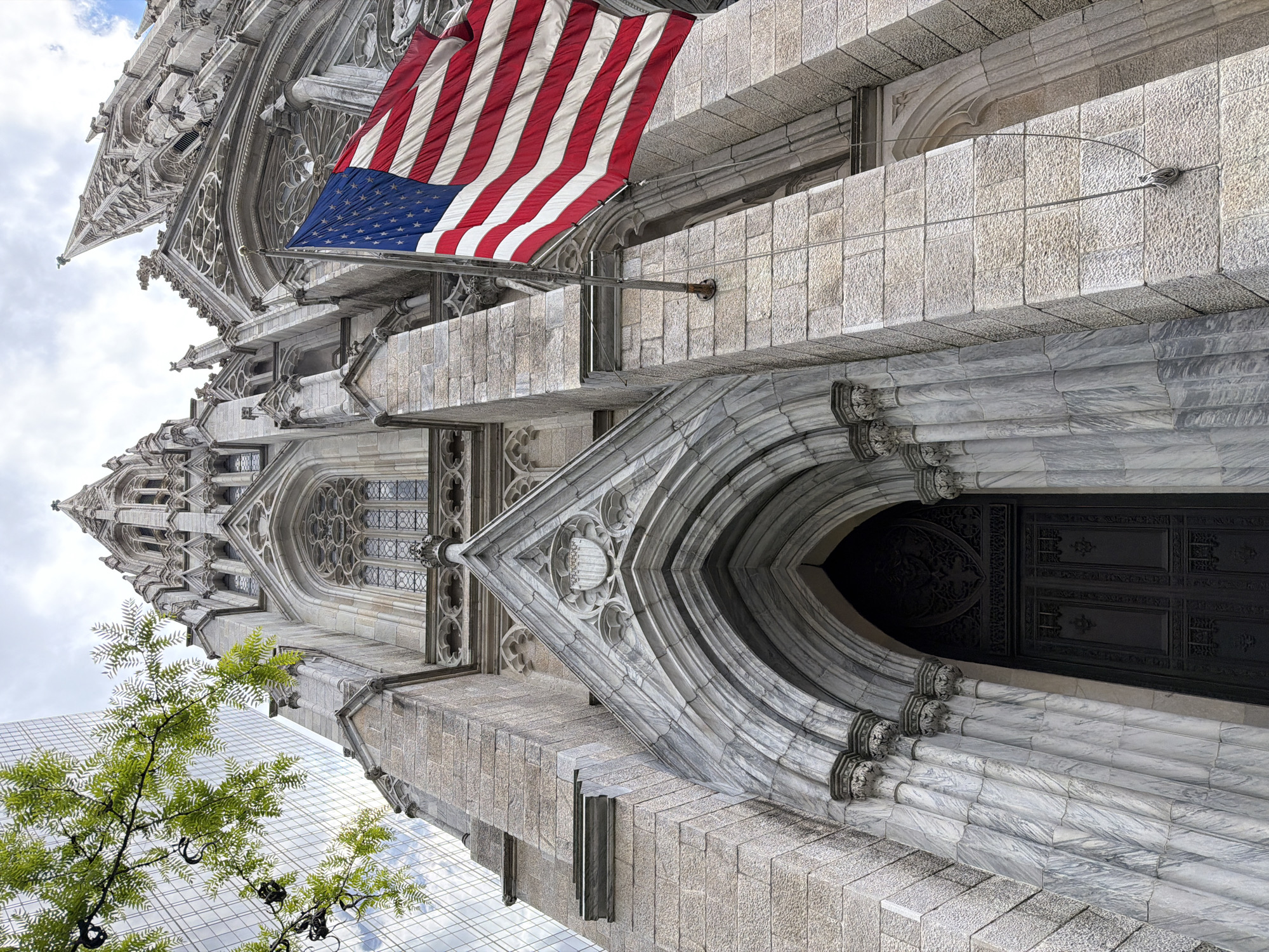 This low-angle shot captures the architectural grandeur of a historical building, adorned with an American flag. The intricate stonework, arched doorway, and towering spires speak to its age and significance. A modern glass skyscraper peeks into the frame, contrasting with the older structure. The presence of a leafy tree adds a touch of nature to the urban landscape. Given the architectural style and the presence of the American flag, this appears to be a photograph of St. Patrick's Cathedral in New York City, taken from a street-level perspective.