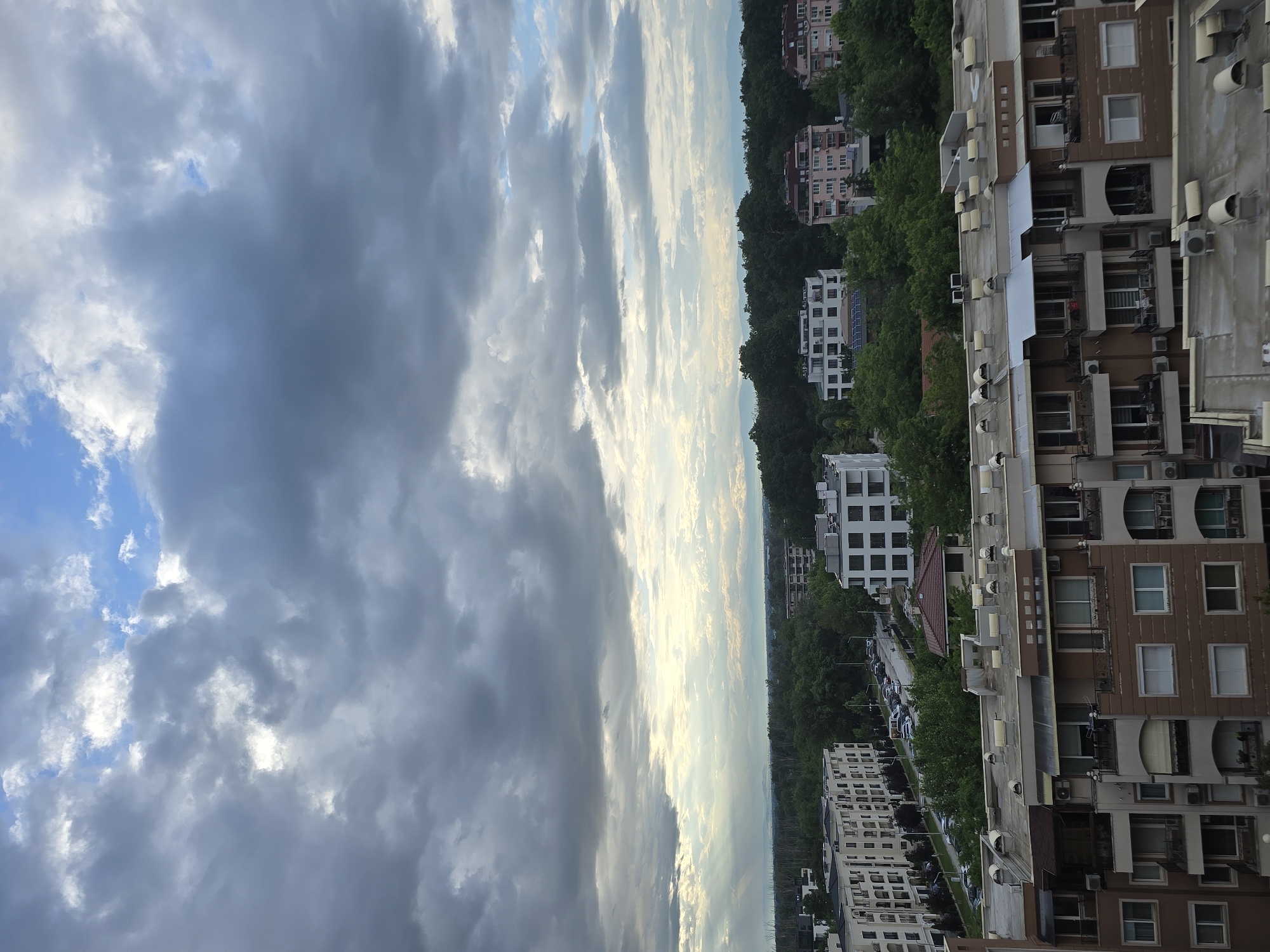 A vertical shot shows a cityscape under a cloudy sky in Unknown, Unknown. The left portion of the frame is dominated by a mix of dark and light clouds with patches of blue sky visible. The right portion displays the cityscape, with a mix of residential buildings and green spaces. The buildings appear to be of varying heights and designs, some with balconies. The presence of air conditioning units on the exterior of the buildings indicates that the climate is warm enough to require them. The image is taken from an elevated viewpoint, possibly from a high-rise building or a hill.