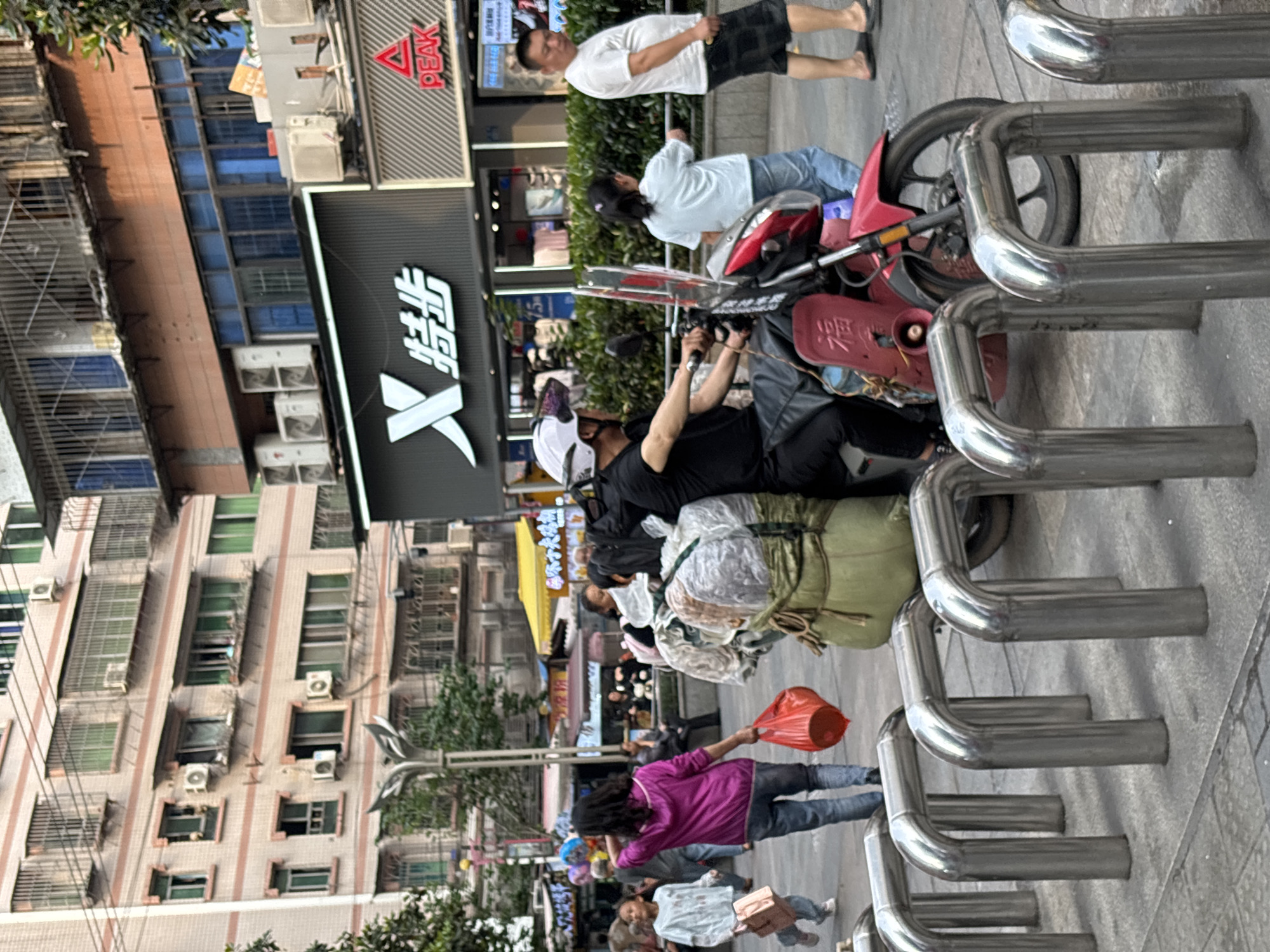 The image depicts a street scene in Jiangjin, China. A row of silver bicycle racks is prominently featured in the foreground. A red scooter is parked behind them. People are walking on the sidewalk. A person on the scooter is wearing a white helmet. Buildings line the street; one features a large sign in Chinese characters and the logo of the brand "PEAK." Air conditioning units are affixed to the buildings' facades. A person carrying a red bag is walking. Bags are tied to the motorcycle.