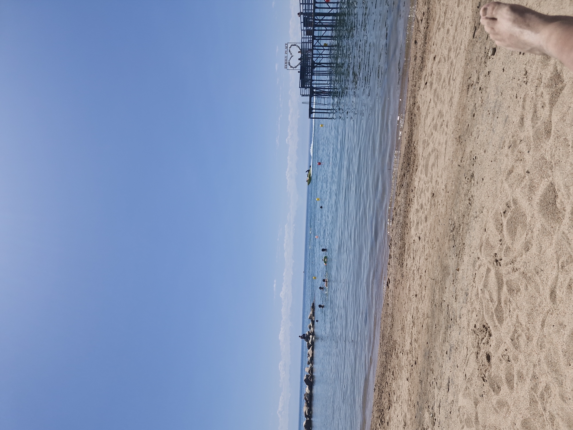 This is a medium-shot, eye-level, outdoor, full-color, vertical photograph taken on a sunny day at a beach. The foreground shows a sandy beach with footprints and a person's feet visible in the bottom right corner. The middle ground depicts a calm, blue body of water with several people swimming and enjoying the water. A breakwater made of large rocks is visible on the left, with a person sitting on top. In the distance, a wooden pier extends into the water, featuring a heart-shaped decoration and a sign that reads "FAMILY PLACE." A yellow jet ski is also seen moving across the water in front of the pier. The background is a clear, bright blue sky with some faint white clouds on the horizon, beyond which mountains are visible. The scene is located in Bulan-Segettu, Kyrgyzstan.