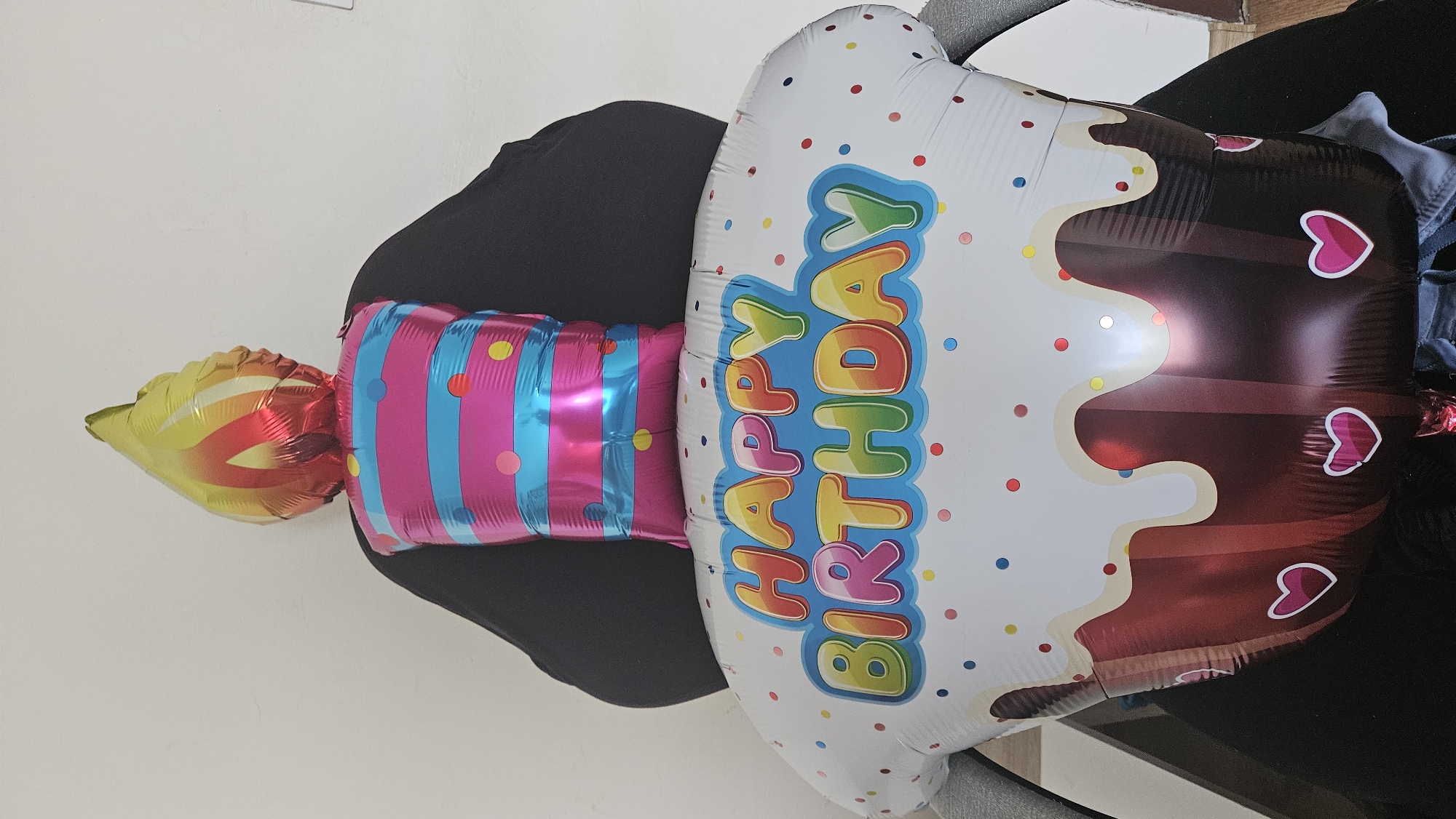 A close-up, eye-level shot captures a large, festive birthday balloon shaped like a cupcake with a candle. The balloon is positioned in front of a plain, white wall. The cupcake base of the balloon is primarily white with an illustration of dripping chocolate frosting and small, colorful confetti dots. The words "HAPPY BIRTHDAY" are prominently displayed in vibrant, rainbow-colored lettering across the top of the cupcake. Above the cupcake is a striped balloon section in pink and blue, topped with a flame-shaped balloon in shades of yellow, orange, and red, resembling a lit candle. The balloons are placed on what appears to be a chair with dark fabric. The background suggests an indoor setting, possibly a home or office, but no specific location in Bratislava, Slovakia, can be identified from the visual information provided. There are no people directly visible, though the balloons are situated on a chair, implying human presence. The lighting is even and appears to be from an artificial source, highlighting the shiny, reflective surfaces of the balloons.