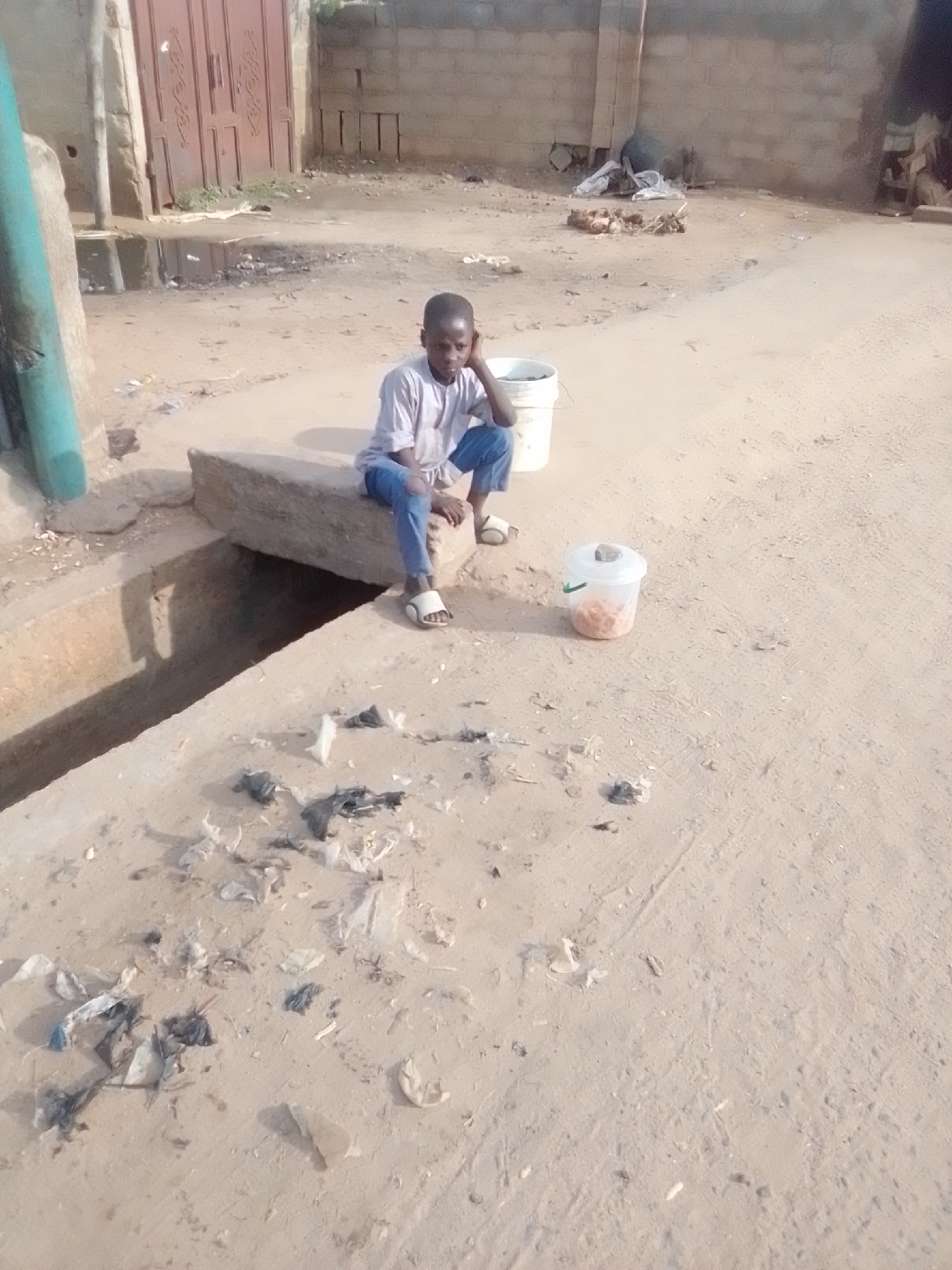 In Galadima, Nigeria, a young boy sits on a concrete ledge next to an open drainage ditch. He is wearing a light-colored shirt and blue jeans, with white sandals on his feet. The boy is resting his head on his hand and looking down. Beside him, a white bucket sits on the ground, and in front of him, a clear plastic container holds a reddish-orange substance. The ground is dirt and sand, scattered with debris, including plastic bags and wrappers. In the background, a red metal gate and a brick wall are visible. The overall scene depicts a somewhat neglected urban environment.