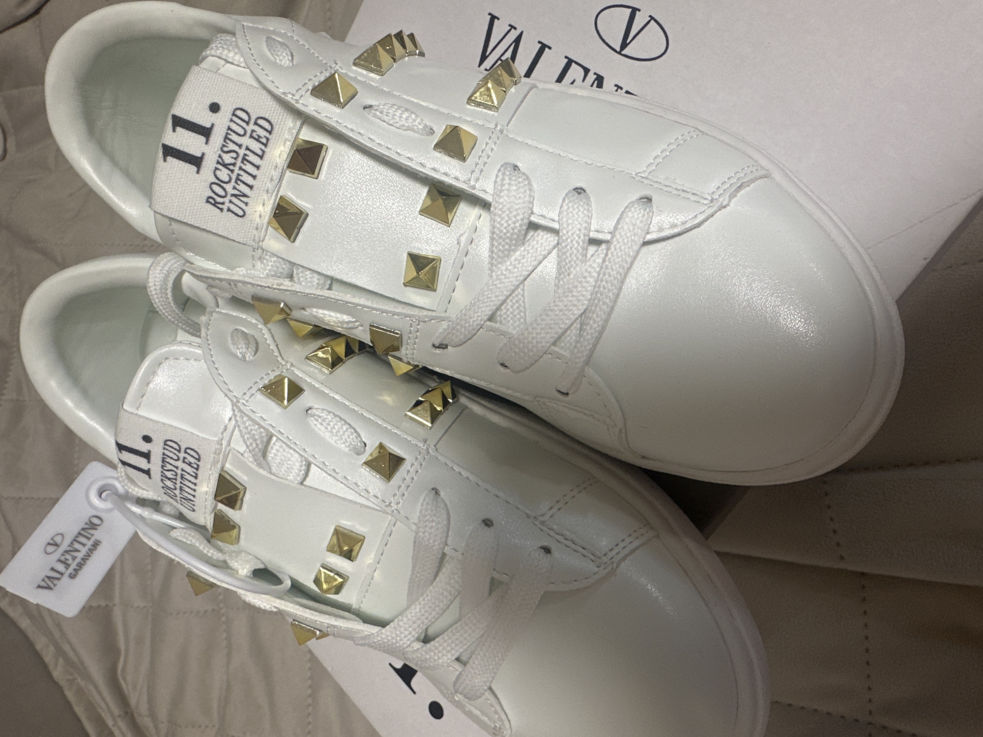 Two white Valentino Garavani "Rockstud Untitled" sneakers are prominently displayed in a top-down, slightly angled view. The sneakers feature a clean, low-top design made of what appears to be smooth white leather. A distinctive design element is the numerous gold-toned, pyramid-shaped studs adorning the sides of the shoes, particularly along the lacing strips and towards the heel. White shoelaces are threaded through eyelets and tied.
On the tongue of each sneaker, a white fabric tag is visible with black text reading "11." and "ROCKSTUD UNTITLED" stacked vertically. A white product tag, bearing the "VALENTINO GARAVANI" brand name and its distinctive "V" logo, hangs from the eyelet of the lower sneaker.
The sneakers rest on a light-colored, possibly padded or quilted surface, with a portion of a white shoe box visible in the background. The box also displays the "VALENTINO" name and its "V" logo in black, confirming the brand. The overall scene suggests an indoor setting, focusing on presenting the new and unworn footwear.