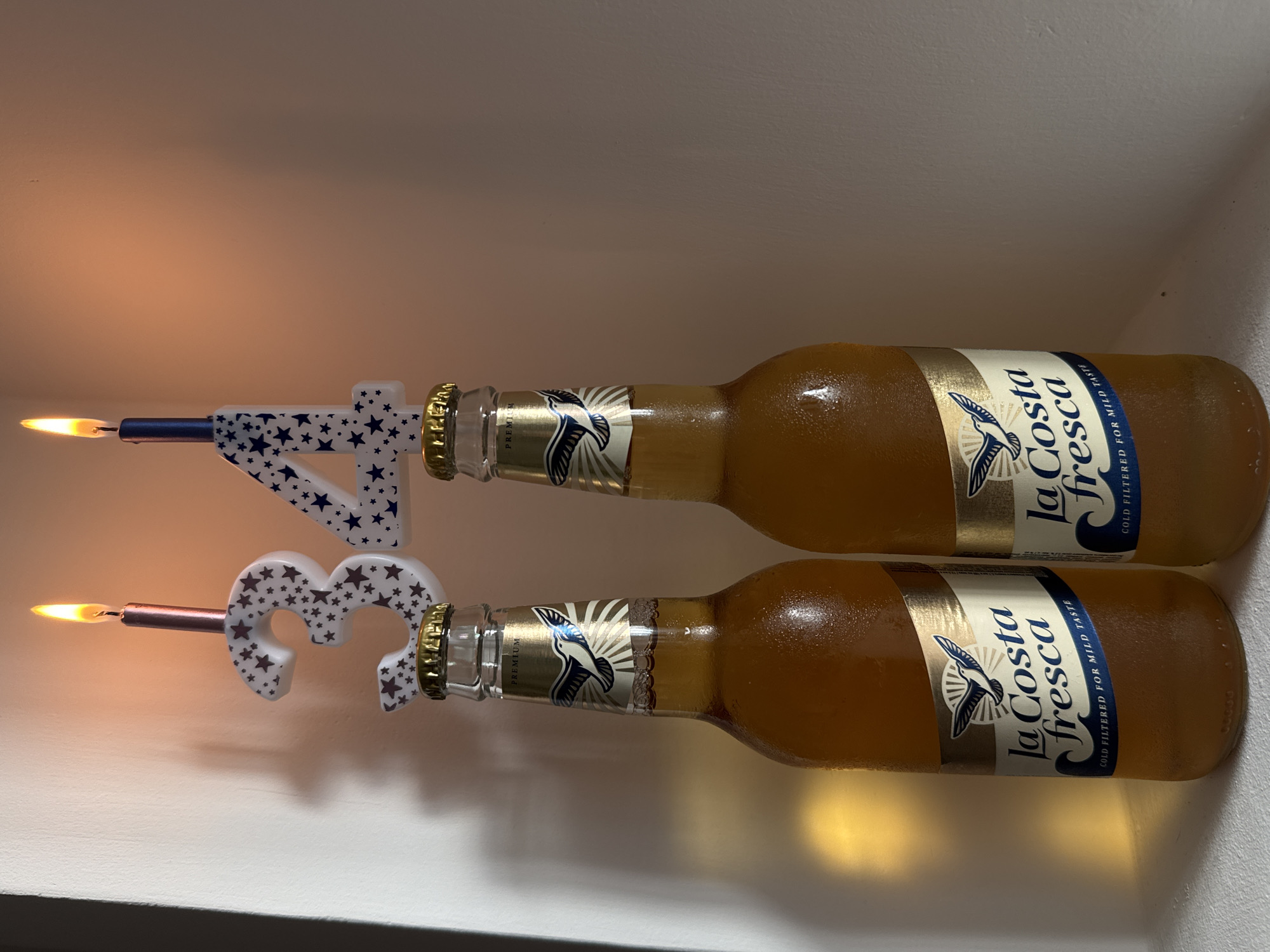 The image displays two condensation-covered glass bottles of "La Costa fresca" beer, presenting a celebratory scene against a plain, light-colored wall in the city of Chaikovsky, Russia.
Each bottle is topped with a numerical birthday candle: a white '3' adorned with dark stars rests on the left bottle, holding a lit rose-gold colored candle, while a white '4' patterned with blue stars sits on the right bottle, holding a lit dark blue colored candle. Both candles are actively burning, producing bright orange-yellow flames and casting warm illumination and soft shadows on the background wall. The glass bottles contain an amber-colored liquid and are covered in fine droplets of condensation, indicating they are cold. Each bottle features a gold-colored cap and a distinctive label: a gold upper band marked "PREMIUM," a stylized bird (resembling a seagull) flying over radiating lines, and the brand name "La Costa fresca" in blue script. Below the brand name, a blue banner reads "COLD FILTERED FOR MILD TASTE." The bottles are resting on a flat, light-colored surface, with a bright, warm light source or reflection visible on the wall to the far left.