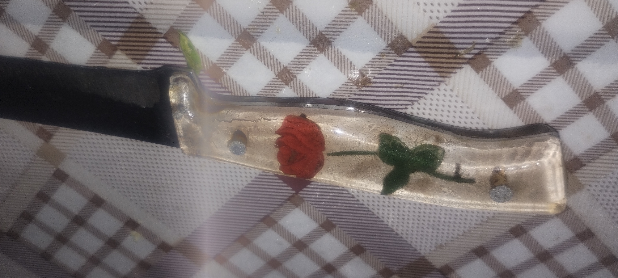 A close-up, slightly angled shot shows the handle of a knife. The handle is made of clear resin with a red rose and green leaves embedded within it. Two metal pins are visible, securing the handle. The blade of the knife, which is black, extends from the right side of the image. The knife rests on a surface covered by a checkered cloth with brown and white squares. The lighting in the image is bright, creating a lens flare across the top of the knife handle.