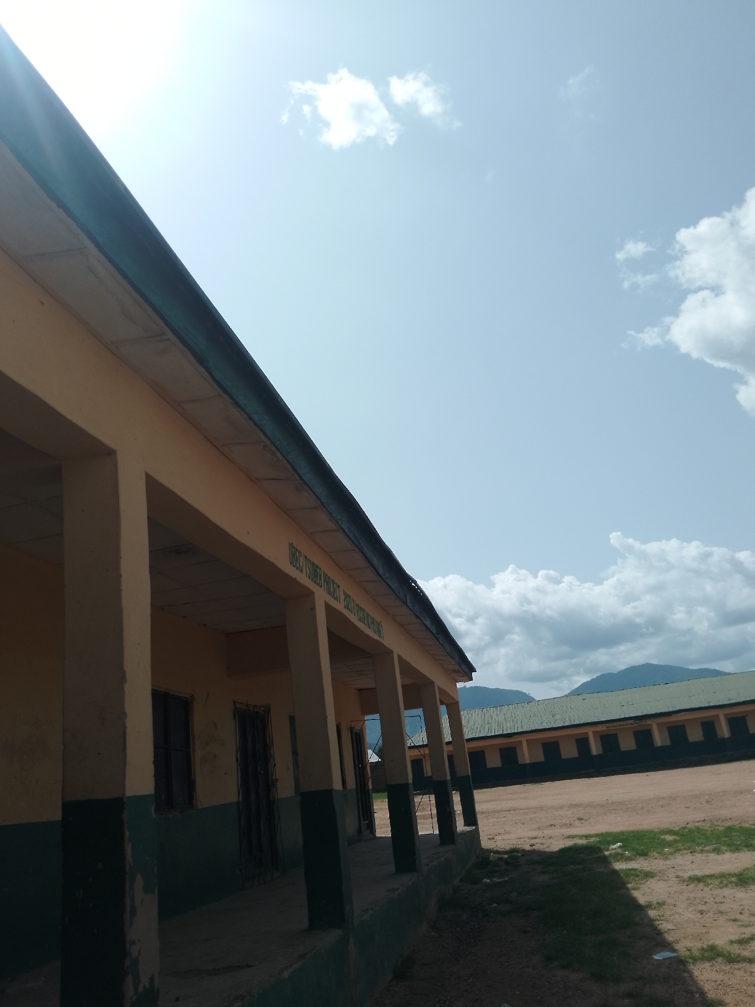 This is a ground-level, eye-level shot of a building in Serti, Nigeria. The building appears to be a school, with a long covered walkway supported by pillars. The walls are painted yellow and a dark green, with some peeling paint visible. Windows and barred doors are visible along the walkway. The sky above is a clear blue with scattered white clouds. In the background, another longer building with a green roof is visible, and beyond that, green hills or mountains can be seen. The ground in front of the building is dirt and patchy grass. The scene is outdoors and appears to be during the day. There are no people or animals visible in the image.