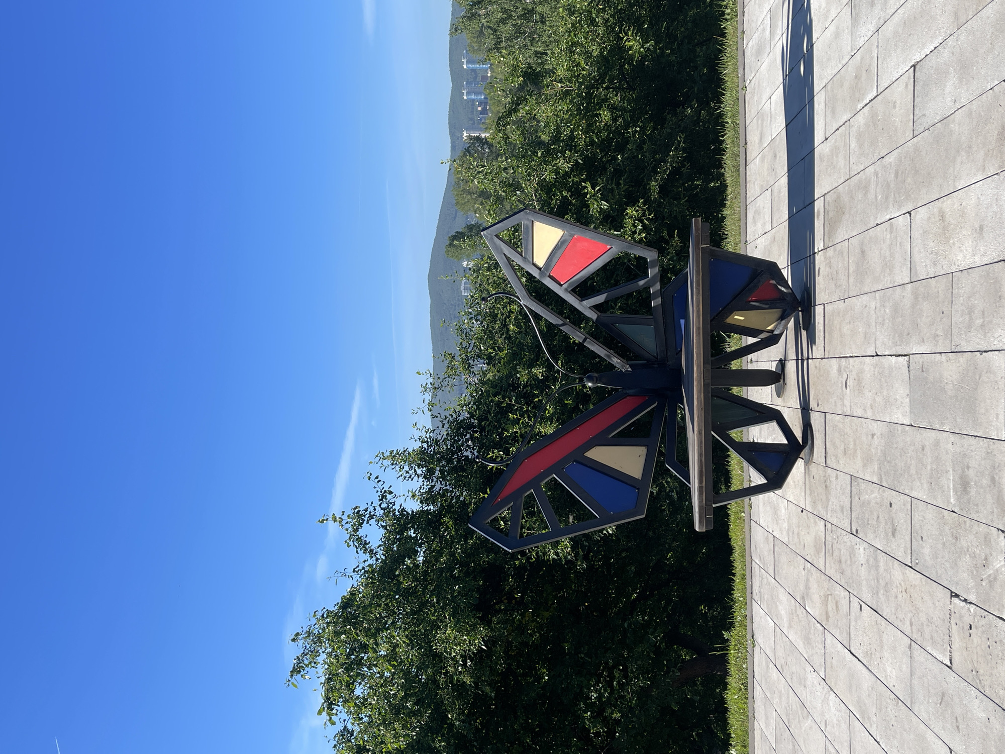 The image depicts a striking, modern metal sculpture shaped like a butterfly, which also functions as a bench. The sculpture is painted with geometric segments of red, blue, and yellow, set against the dark metal frame. It is positioned on a paved walkway made of light-colored square tiles, with the butterfly facing the viewer. Behind the sculpture, dense green trees form a natural backdrop, with a distant view of a city skyline featuring modern buildings nestled among hills under a clear, bright blue sky. The scene is bathed in sunlight, casting distinct shadows on the paved area. The sculpture is located at the observation deck on Grigoriev Street in Krasnoyarsk, Russia, a popular spot offering panoramic views of the city and the Yenisei River. There are no people visible in the image.