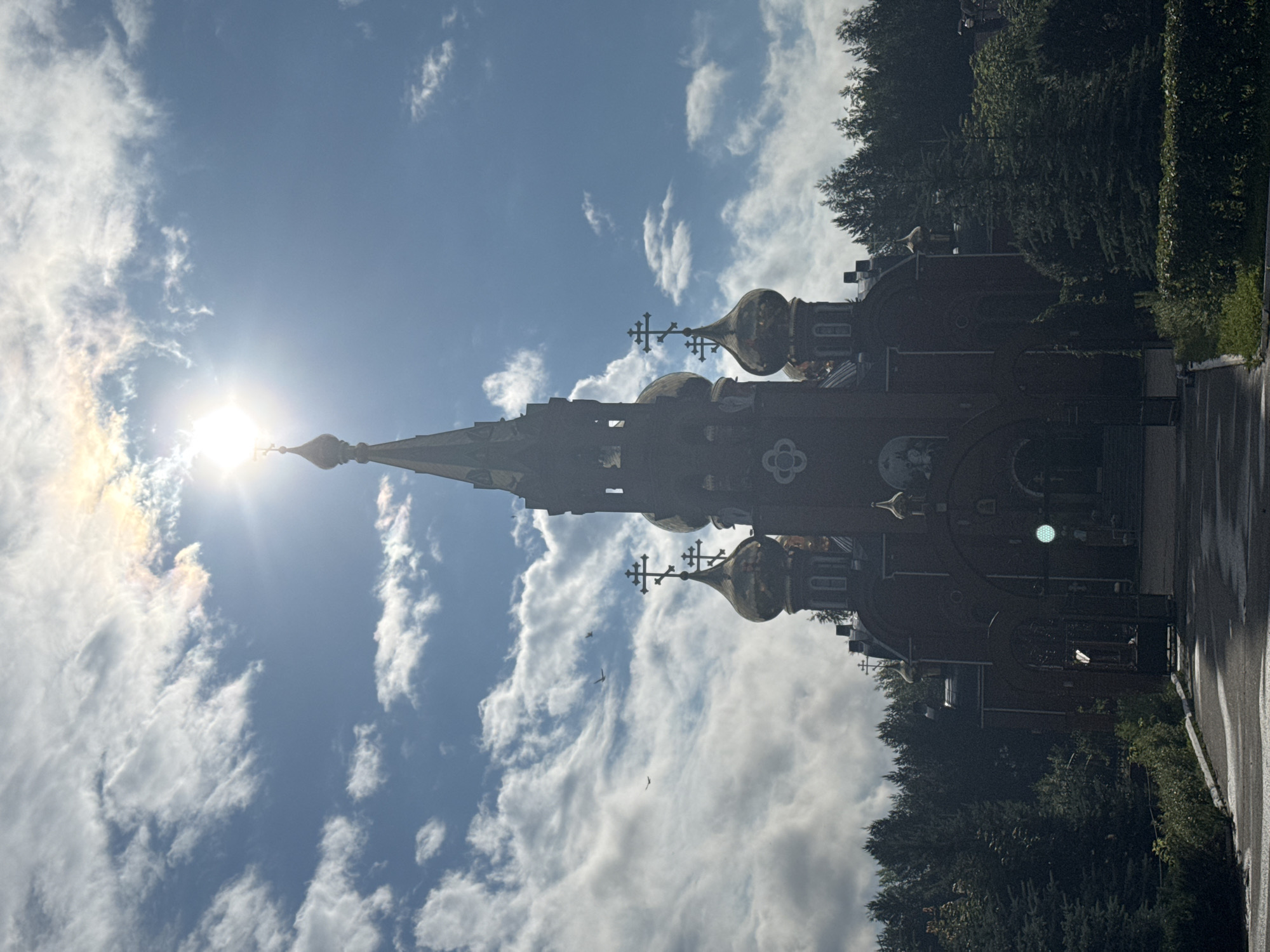 The image shows a view of the Church of the Assumption of the Blessed Virgin Mary in the city of Chaikovsky, Russia. The church, constructed of red brick, features multiple golden domes and a tall, spire-like central tower topped with a cross. The sun is bright and partially obscured by clouds, creating a lens flare effect. The church is surrounded by lush green trees, including pine trees. A paved pathway leads up to the church entrance, which is partially visible behind a dark metal gate. The sky is blue with scattered white clouds, and several birds are in flight. The overall scene is serene and majestic, highlighting the architectural beauty of the church against a natural backdrop under a bright sky.