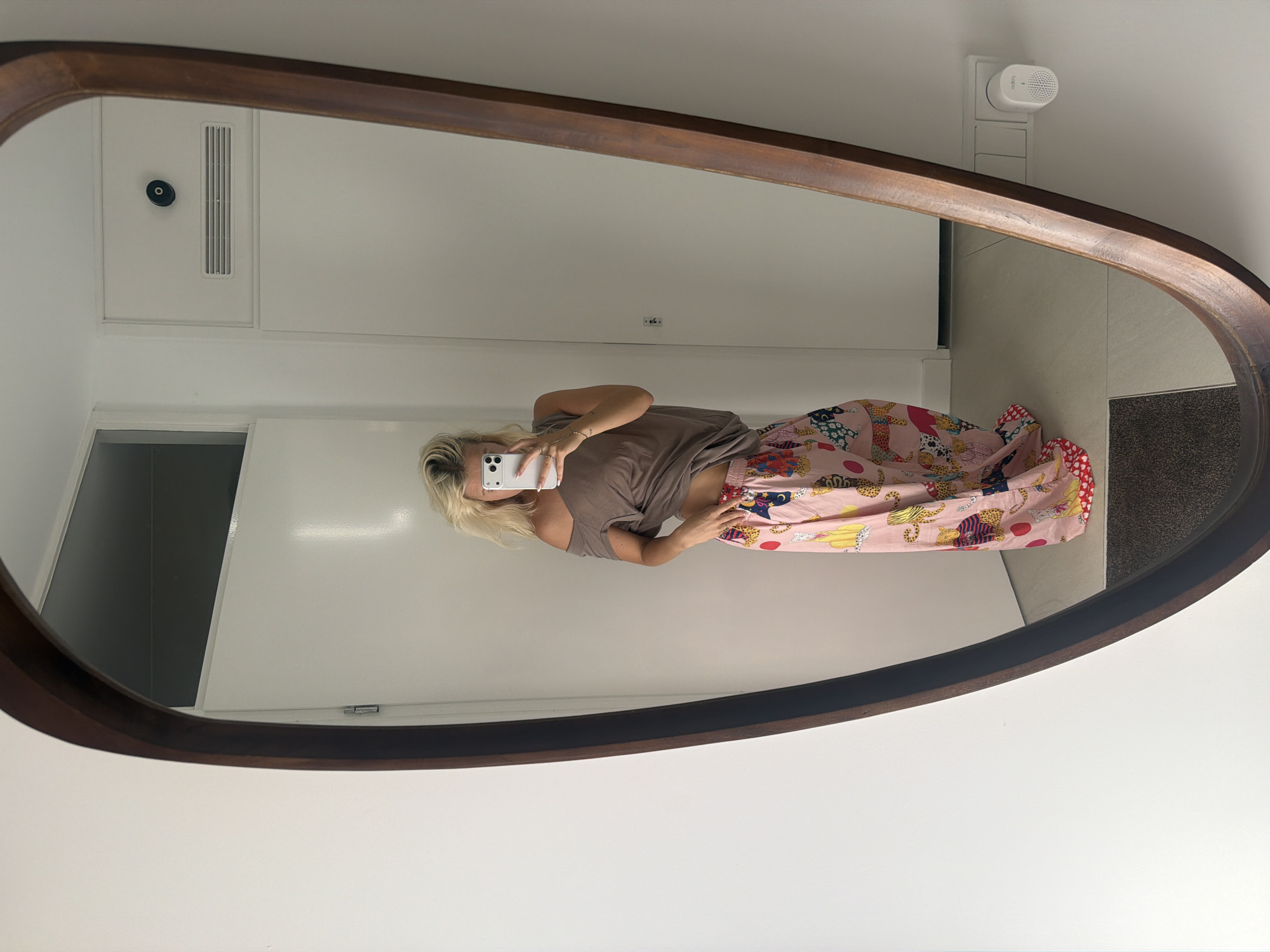 A blonde woman, reflected in a large, oval, dark wood-framed mirror, stands in a well-lit, modern interior. She is wearing a taupe, off-the-shoulder top and pink, wide-leg pajama pants adorned with a colorful pattern of leopards, moons, and other whimsical figures. Her hair is styled in a messy bun. She holds a smartphone in her right hand, taking a selfie, and her left hand rests on her hip. The background of the reflection reveals a white wall with a closed door, a ventilation grille, and a white electrical outlet with a small white device plugged into it. The floor appears to be tiled with beige tiles. The overall impression is one of a casual, domestic setting. This image was captured in a home in Ridderkerk, Netherlands.