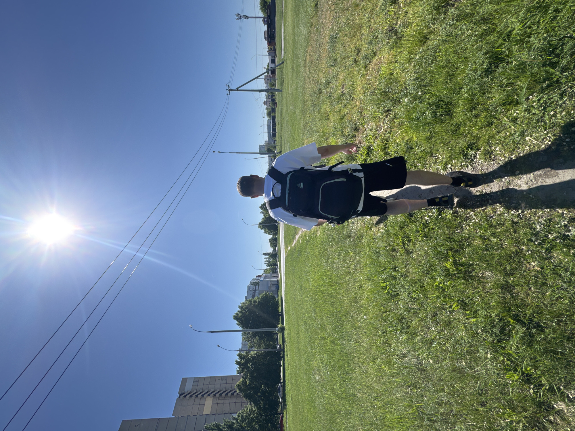 A man wearing a white shirt and black shorts is walking along a sidewalk bordered by a grassy area. He has a black backpack on. The sun is shining brightly in a clear blue sky. Utility poles and lines are visible in the background. In the distance are buildings and trees, suggesting an urban or suburban environment.