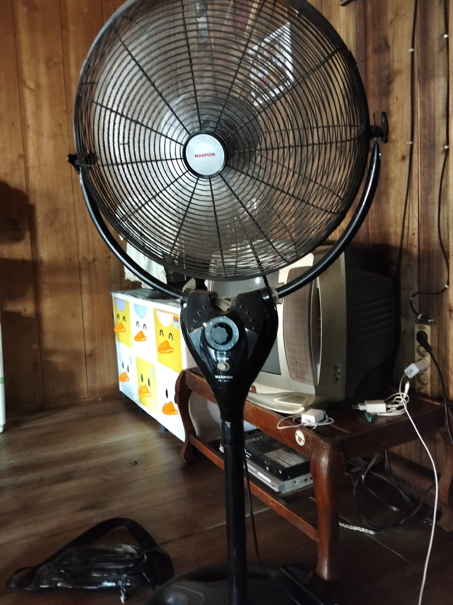This image depicts the interior of a room, likely a residence, in Setia Jaya, Indonesia. The primary subject is a tall, black electric fan with a large, circular grille. The fan is positioned centrally and prominently in the frame. To the right of the fan, a vintage television set with a boxy CRT screen is visible, placed on a wooden stand. Various electrical cords and adapters are scattered around the television and the stand, suggesting a functional, though somewhat cluttered, electronic setup. Behind the fan and television, a wall paneled with wood planks creates a rustic background. In the lower left quadrant, a black sling bag or waist pack lies on the wooden floor. A small cabinet decorated with cartoon duck faces is positioned to the left of the television. The lighting in the scene appears to be ambient, with shadows cast by the fan and other objects. The overall impression is one of a lived-in space, possibly a living room or study area, with everyday objects arranged around the fan and television.