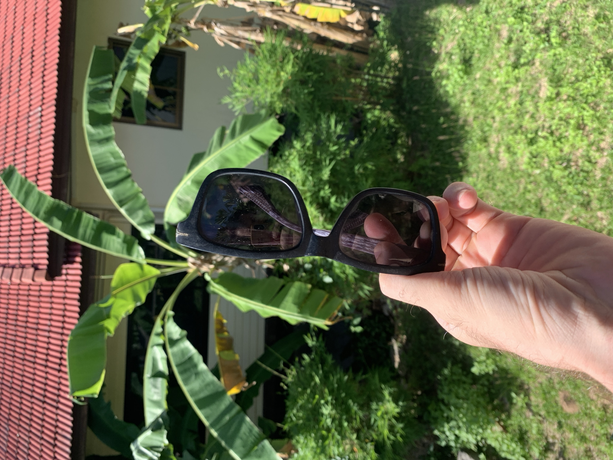 A person holds a pair of black sunglasses in their right hand. The reflection in the lenses reveals an outdoor scene. A building with a red tiled roof and white walls stands to the left, partially obscured by large, green banana leaves. In the background, lush green foliage and grassy areas are visible. The location is outdoors in บ้านบางรักษ์, Thailand, as deduced from the surrounding vegetation and architectural style.
