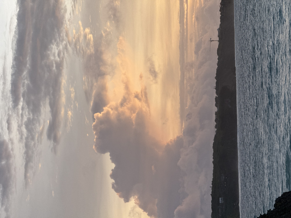 The image captures a coastal landscape at dusk or dawn in Pecatu, Indonesia. The upper two-thirds of the frame are filled with a complex cloud formation. Large, voluminous cumulus-type clouds, illuminated with orange, pink, and yellow light from the right, dominate the mid-sky, transitioning to darker grey and purple tones on their left. Above, thinner, streaky grey clouds are visible. The horizon is marked by a dark, silhouetted landmass, appearing as a tree-covered cliff or peninsula. On this landmass, indistinct white structures are present on the left side, and a prominent yellow and white construction crane is clearly visible on the right. The lower third of the image depicts a body of water, likely the ocean, with discernible wave patterns and a dark blue-green color. A dark, jagged rock formation is partially visible in the bottom-left foreground. No individuals, specific actions, or direct interactions are visible.
