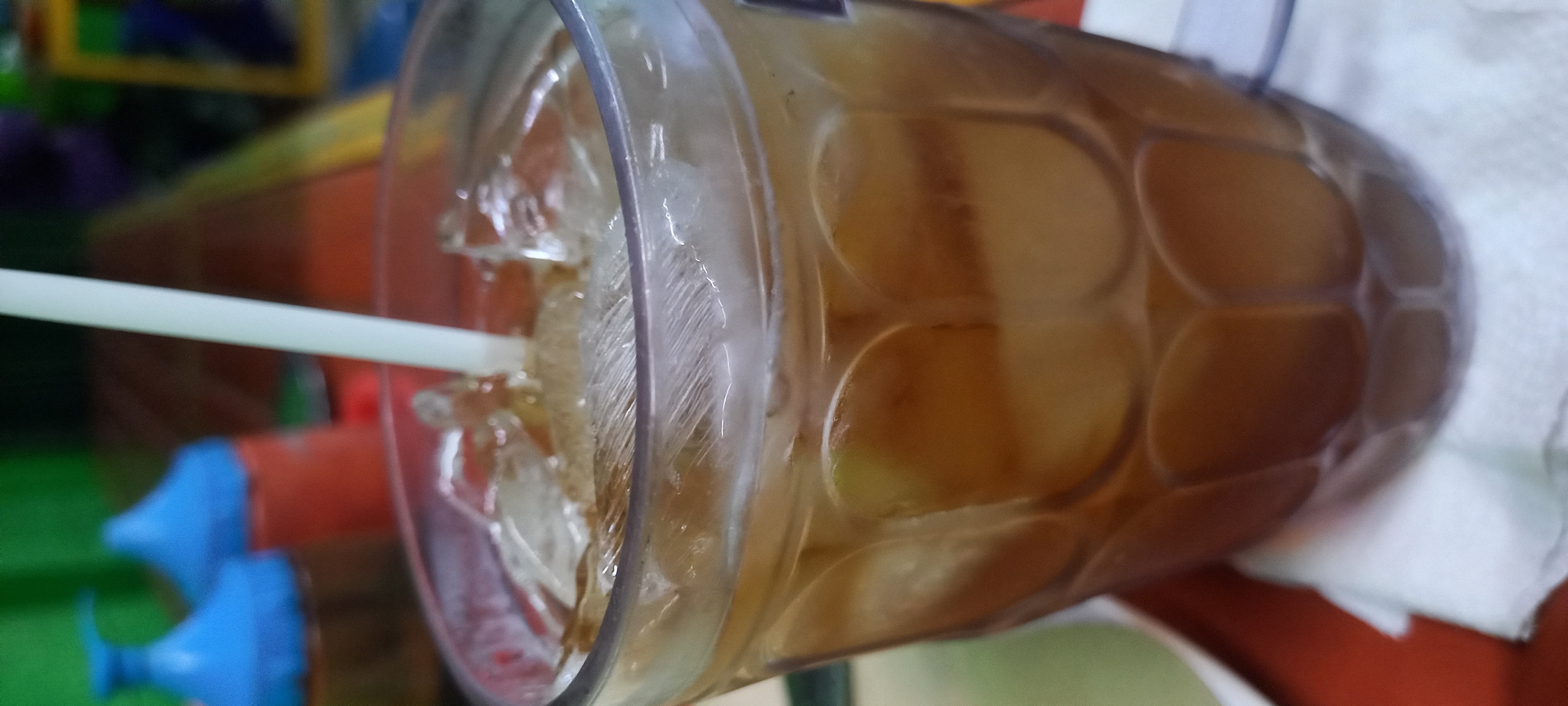 An iced tea drink sits on a white napkin, which is placed on a red surface. The drink is in a clear glass filled with ice cubes and a pale yellow liquid, with a white straw submerged in it. In the background, out of focus, are two orange containers with blue lids and a green surface. The scene appears to be outdoors in Loa Lepu, Indonesia, likely at a food stall or eatery given the context of the items present.