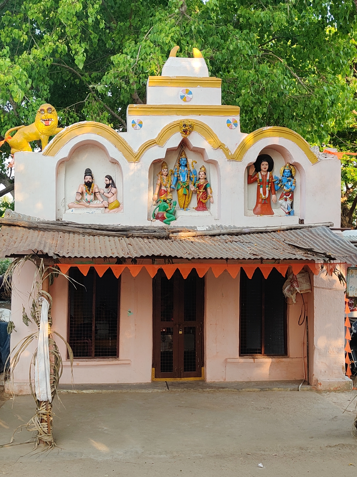 This image displays a brightly adorned Hindu temple or shrine located in Mamillakunta, India, likely captured during the warm, soft light of the golden hour. The structure is predominantly light pink with a corrugated metal roof and a white upper facade featuring golden arched borders and colorful circular patterns.
The upper part of the temple showcases three prominent niches containing colorful statues. In the leftmost niche, two bearded male figures, possibly rishis or sages, are depicted in seated or meditative postures. The central and largest niche features a depiction strongly suggesting the Rama Darbar, with a blue-skinned figure (likely Lord Rama) holding a bow, accompanied by a female figure (Sita) and two smaller figures (potentially Lakshman and Hanuman or Garuda). The rightmost niche holds a statue of a figure in red robes with dark, curly hair, possibly a revered saint like Sai Baba, standing with an uplifted hand, alongside another blue-skinned deity, likely Krishna or Vishnu.
Perched on the left side of the roof's upper tier is a vibrant yellow, stylized lion statue with an open mouth and bared teeth.
Below the deities, the main temple structure has a simple light pink facade with a central wooden door and two barred windows, all appearing closed. An orange triangular bunting banner stretches across the front below the roofline. To the left of the entrance, a pole wrapped in dried leaves and a white cloth stands upright. The ground in front is a simple dirt or concrete surface. In the background, a large, lush green tree provides a natural canopy. On the right side of the building, a partially visible white sack or bag with some obscured text (possibly "WAK CEM") is hanging. The overall scene conveys a sense of devotional calm and cultural richness.