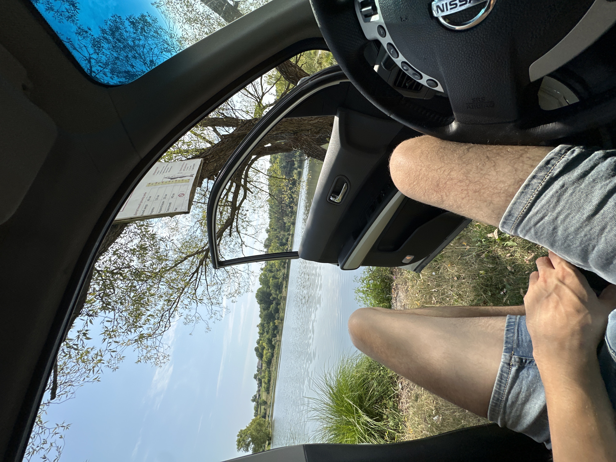 The image captures a view from inside a car, parked near a body of water. The driver's side door is open, revealing a serene landscape of a lake, green vegetation, and trees under a clear blue sky with some wispy clouds. A sign is visible on the windshield, which appears to be in Ukrainian, detailing fishing regulations. The focus is on the car interior, with the steering wheel and dashboard in the foreground, and the legs of a person wearing denim shorts visible. The location is identified as the city of Makiv, Ukraine, suggesting a scenic spot for recreation.