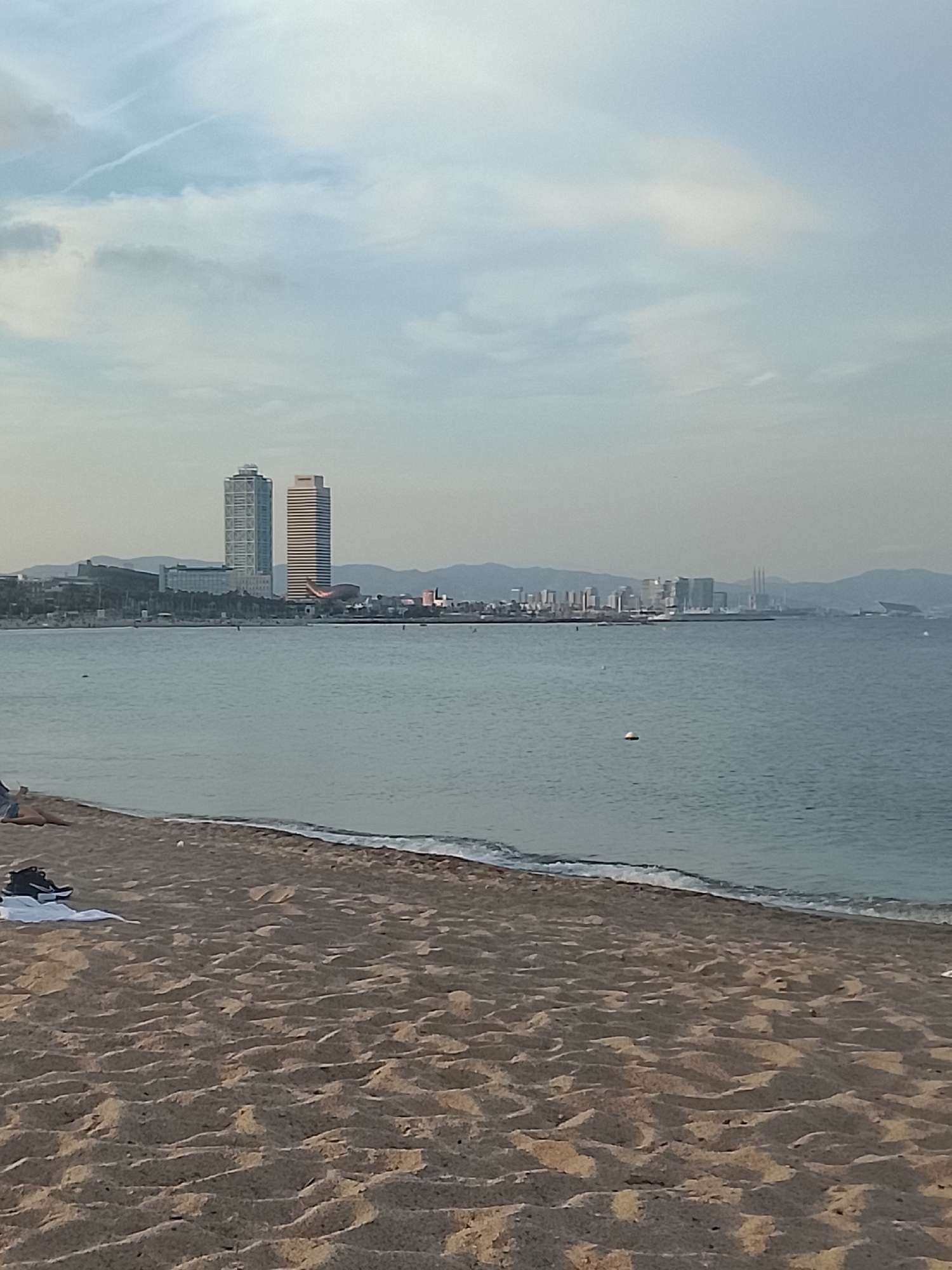 The image captures a panoramic view of a beach in Barcelona, Spain, looking out over the Mediterranean Sea towards the city skyline and distant mountains under a pale, hazy sky.
In the foreground, the sandy beach displays subtle ripples and texture, with a thin line of gentle waves breaking at the water's edge. A white towel and a pair of dark shoes are visible on the sand in the lower-left corner. A person or two can be faintly discerned lying down on the sand further to the left, partially cropped out of the frame.
The middle ground features the calm, greyish-blue expanse of the sea, reflecting the muted light from the sky. Several small, white buoys are scattered across the water.
Across the water, the iconic skyline of Barcelona is prominent. Two tall, distinct skyscrapers, the Hotel Arts and Torre Mapfre, stand out. To their right, a collection of other city buildings and industrial structures, including power station smokestacks, stretch along the coastline. Faint outlines of mountains rise in the far background.
The sky is a light, overcast blue with some wispy clouds and a few faint contrails. The soft, diffused lighting suggests it is late afternoon or early evening. The scene conveys a tranquil and relaxed atmosphere. No visible text is present in the image.