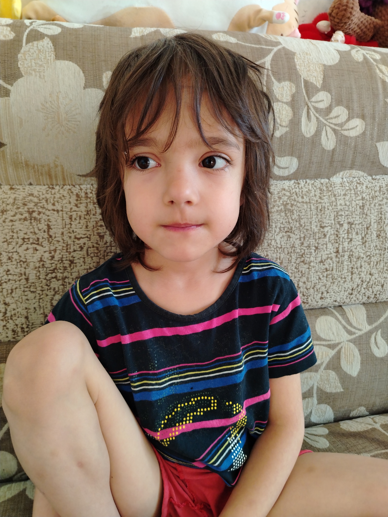 A young child with dark hair and brown eyes is sitting on a couch. The child is wearing a black t-shirt with horizontal stripes of pink, blue, yellow, and white, and red shorts. The couch is upholstered in a patterned fabric with floral designs. In the background, on the couch, are several stuffed animals, including a tan and white rabbit, a red stuffed animal, and a brown crocheted bear. The image was taken indoors. No specific location in the city of Noviy, Russia can be determined from the image.