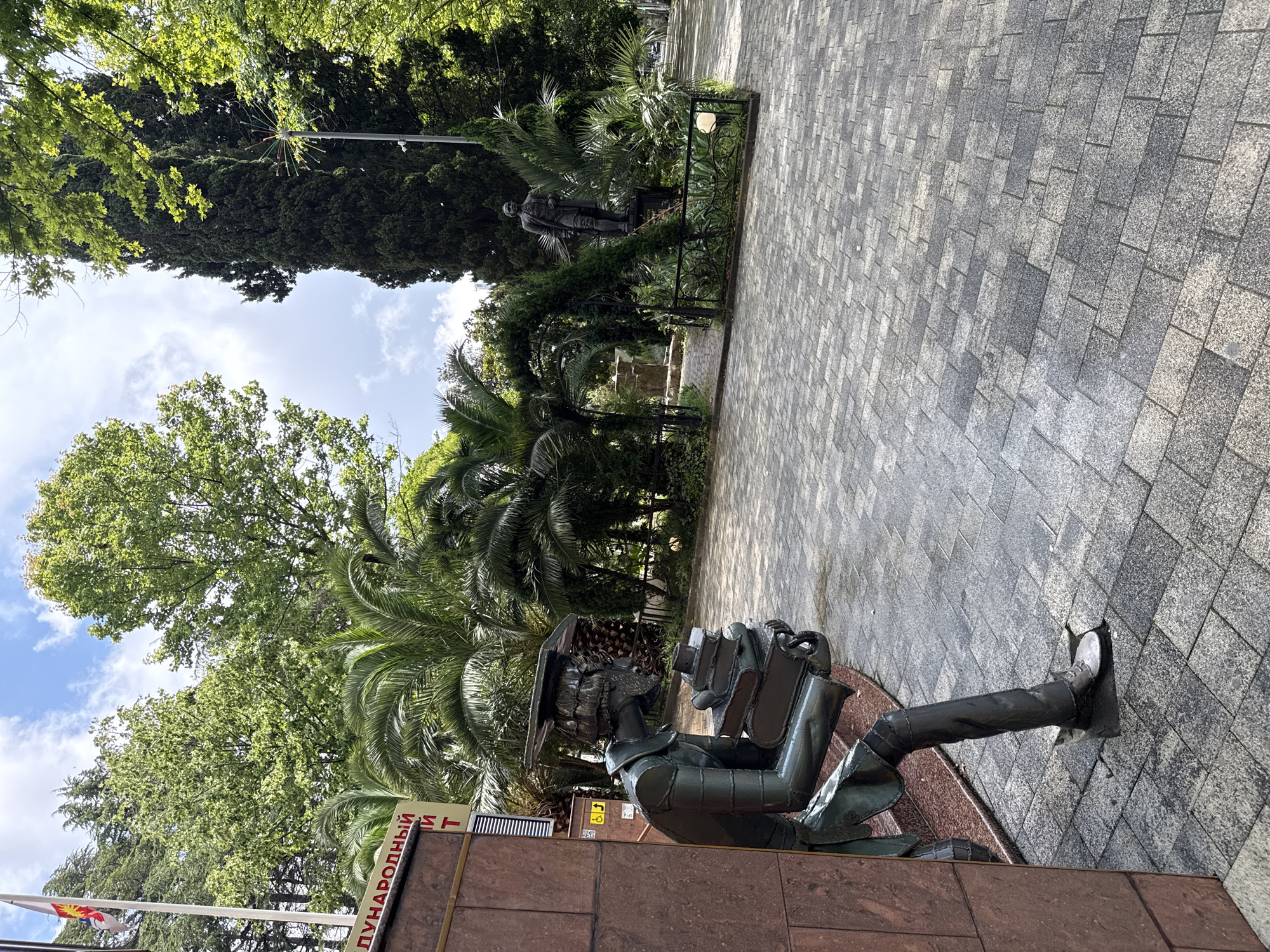 In Sochi, Russia, a sculpture of a scholar with a mortarboard hat and a stack of books is positioned near a textured brown wall. The scholar, made of dark metal, is depicted in a seated pose, leaning against the wall and clutching books. In the background, a paved walkway leads to a statue of a man in historical attire standing behind a decorative fence. Lush green trees, including palm trees and a tall cypress tree, surround the area. A flag with a sun and star emblem is visible on a flagpole to the left. The sky is partly cloudy, with a bright, clear day evident.