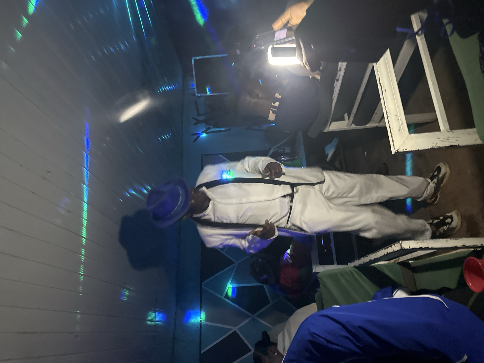 The image captures a man dressed in a white suit and fedora dancing indoors, illuminated by blue and green lights that cast reflections on the ceiling. He is standing in the center of the frame, with his body angled slightly to the right as he dances. To his left, several people are seated, some partially obscured. One person in the foreground to the left wears a blue jacket. To the right, another individual is holding up a phone, seemingly recording the scene. The environment appears to be a dimly lit room with a patterned wall in the background and seating arrangements. The scene is set in Wumat, Nigeria.
