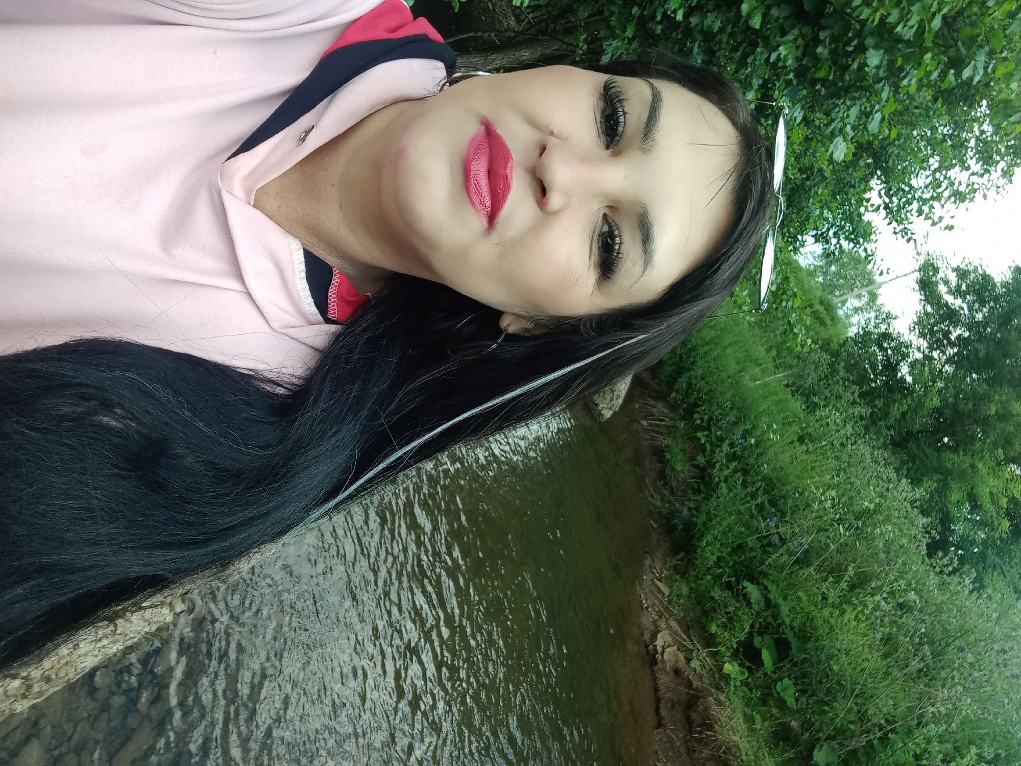The media file is a selfie of a woman with long, dark hair and bright red lipstick. She is wearing a pink hoodie with navy blue and red accents. She is looking at the camera with her head tilted. Behind her is a body of water with a rocky edge, and lush green trees and vegetation. There are no visible text or specific details about the time of day or weather, but the lighting suggests it is daytime. The overall mood seems relaxed and casual. The setting appears to be a natural outdoor environment, possibly a park or a riverbank.