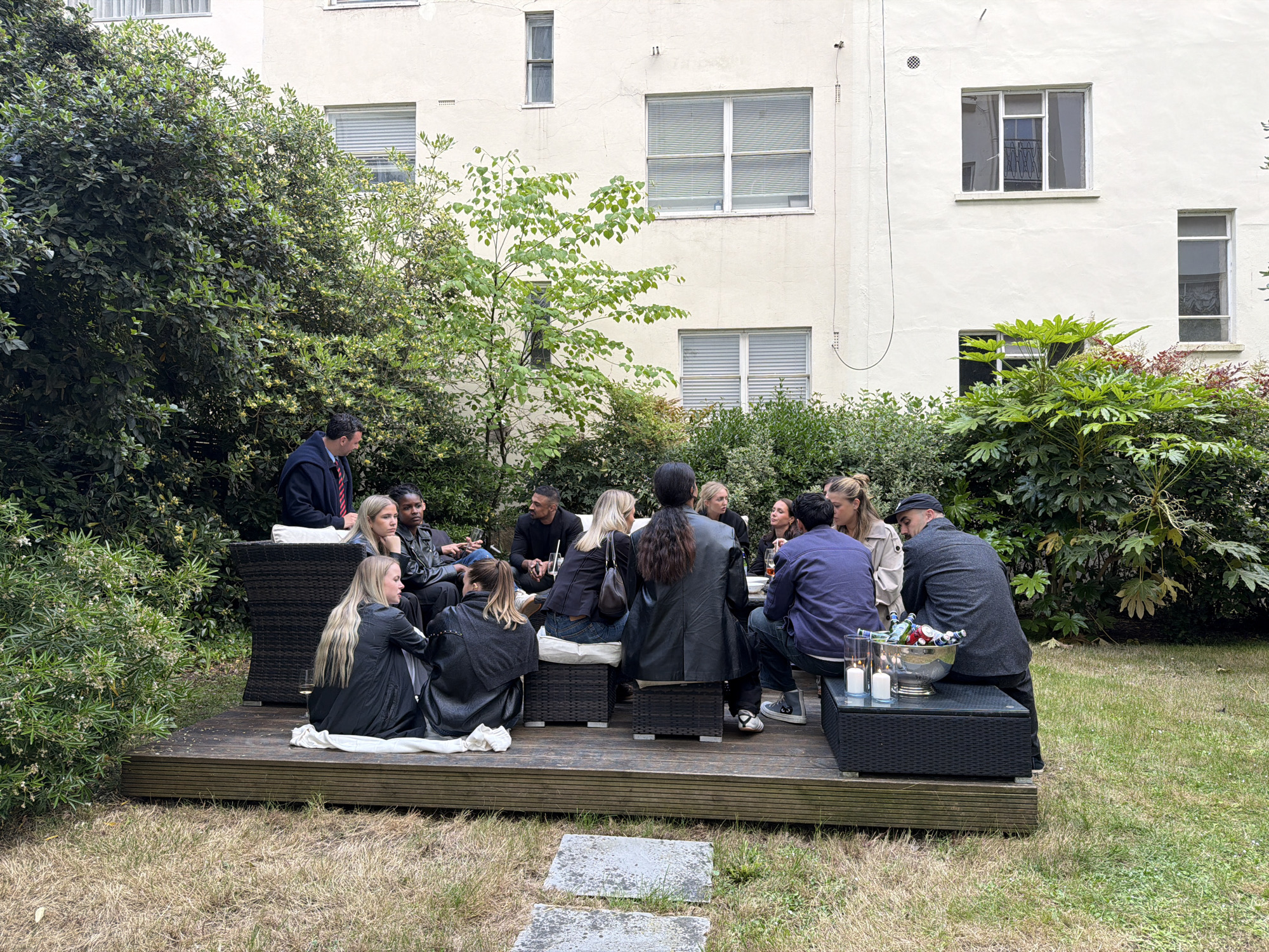 A group of approximately 14 people are gathered on a wooden deck in an outdoor garden setting, surrounded by greenery, with a light-colored multi-story building in the background. The scene appears to be in London, United Kingdom, based on the architectural style and environment.
The people are of various ethnicities and are casually dressed. They are sitting and standing around a central area with low tables and seating arrangements made of dark wicker-like material. Some are engaged in conversation, while others are looking in different directions. There are drinks and candles on the tables.
The deck is slightly elevated from the grassy lawn that surrounds it. The building behind them has multiple windows of varying sizes. The scene is bathed in natural light, suggesting it is daytime.