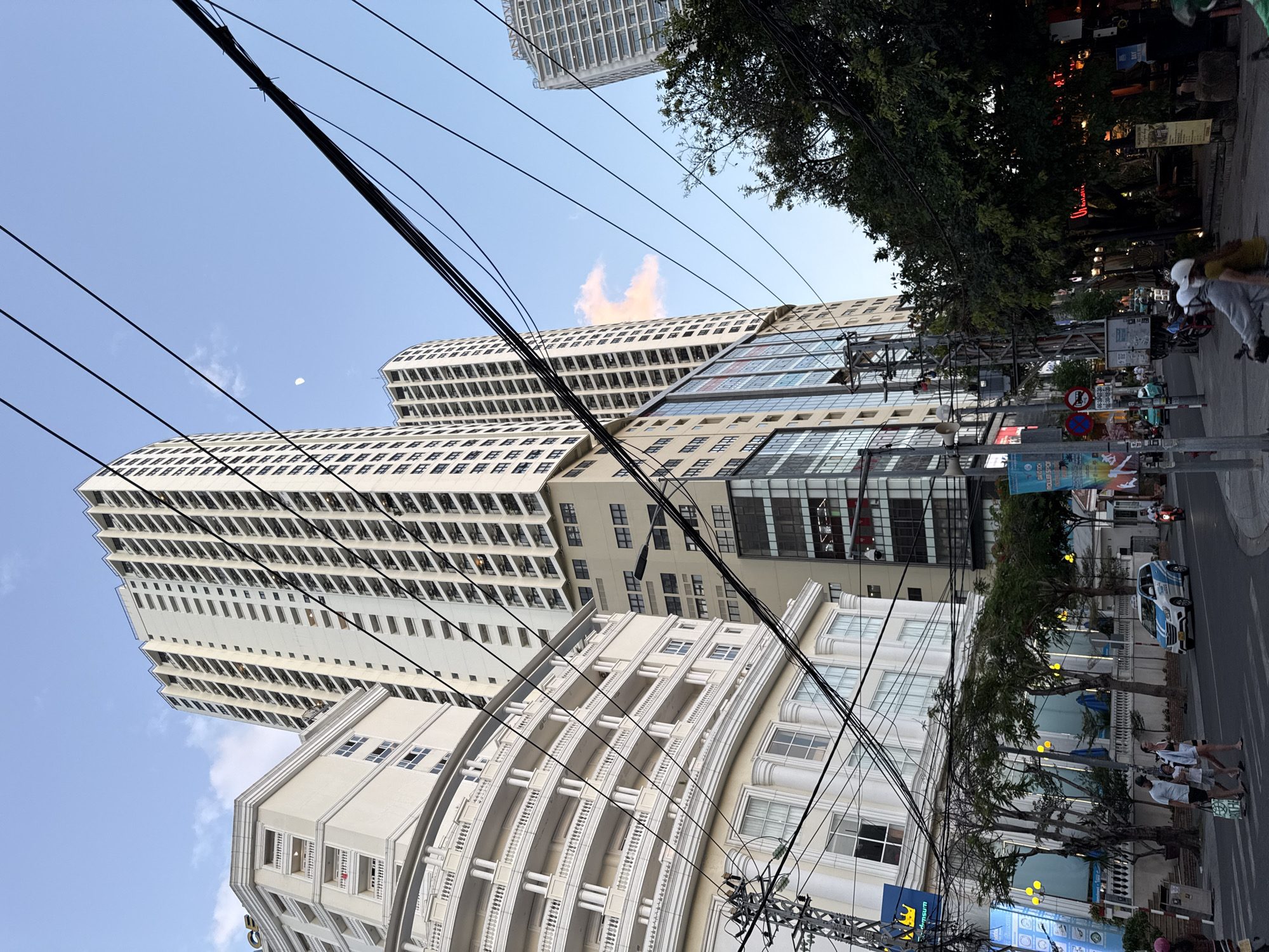 The image shows a street view in Nha Trang, Vietnam, characterized by tall apartment buildings, overhead power lines, and pedestrian activity. In the foreground, a street scene unfolds with people walking across a crosswalk and a white and blue car driving on the road. Several trees line the sidewalk, providing some greenery amidst the urban landscape.
Dominating the background are multiple high-rise buildings. To the left, a curved, ornate building with balconies and columns suggests a hotel or residential complex. Behind it, a very tall, slender white apartment building stretches towards the sky, its facade dotted with numerous windows. Further back, another similarly tall building with a grid-like pattern of windows is visible. A smaller building with a more modern glass facade is situated between the curved building and the taller apartment buildings. The sky is partly cloudy with a hint of sunset colors and a sliver of the moon is visible. A variety of businesses, including shops and restaurants, are indicated by signage and illuminated windows along the street. The presence of numerous power lines crisscrossing the frame is a prominent feature of the urban environment.
