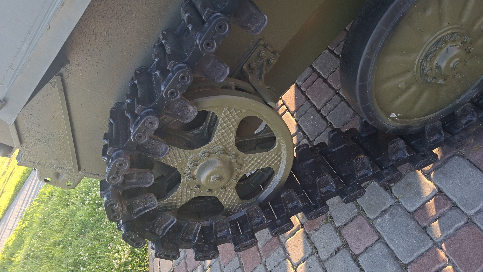 The image shows a close-up view of a tank's tread and a portion of its hull, resting on a paved surface made of interlocking bricks. The tank is painted in a military green color. The tread is made of black metal links with visible bolts and pins. A large, olive-drab sprocket wheel with a starburst pattern is prominent, engaging the tread. To the right of the sprocket, another olive-drab road wheel with a ribbed tire is visible. The background includes a strip of green grass with small white flowers and a paved path. The lighting suggests it is daytime, with sunlight casting shadows. The scene is static, with no people or overt actions depicted. The location is identified as the city of Smolevichi, Belarus.