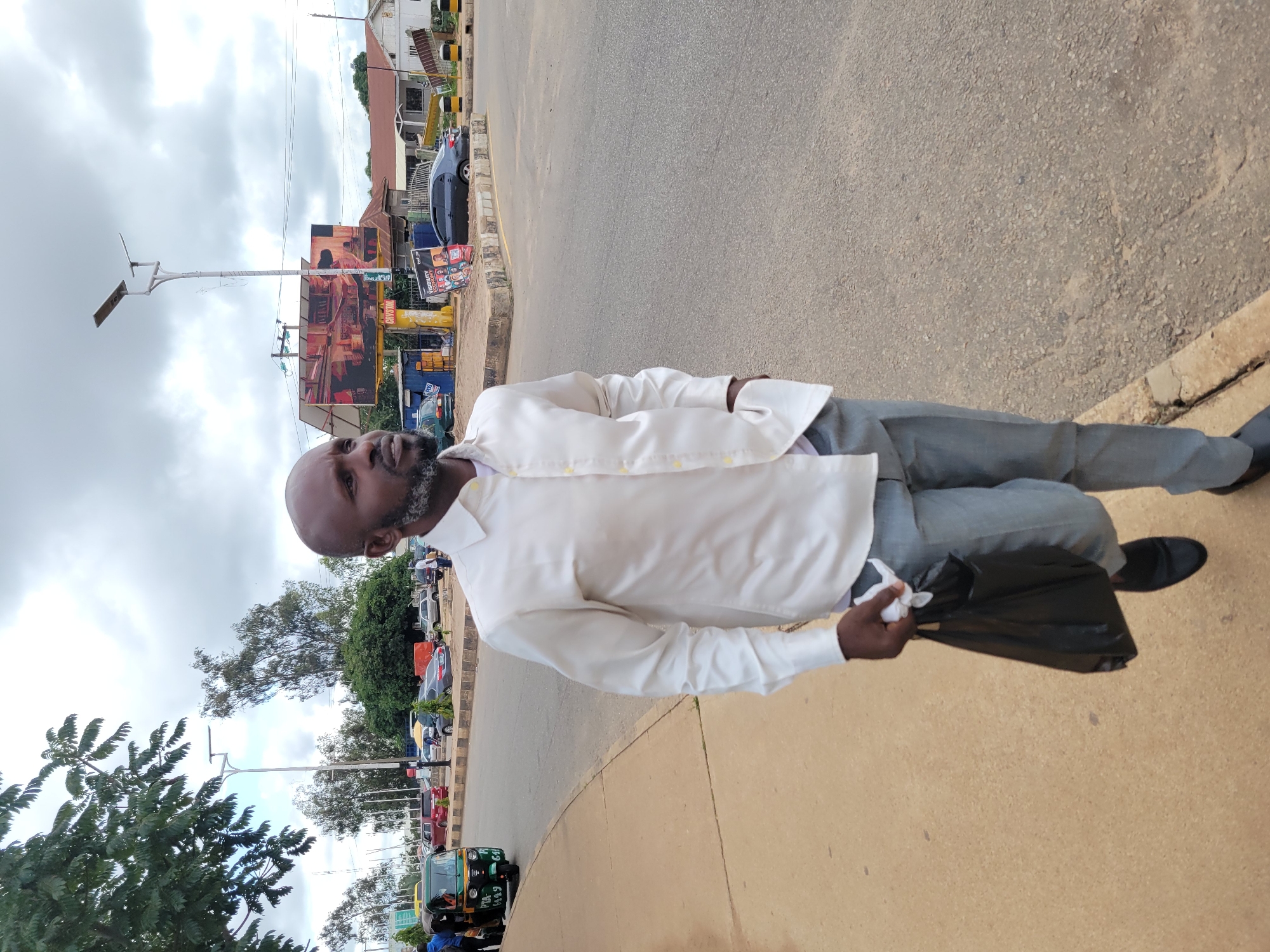 A man with a bald head and a grey beard walks along a sidewalk in Jos, Nigeria, holding a black plastic bag in his right hand. He is wearing a long-sleeved white collared shirt and light grey trousers, with one hand in his pocket. The man is walking away from the camera, looking to his left. In the background, a busy street is visible with several cars, a three-wheeled auto-rickshaw, and buildings. A large billboard stands prominently on the right side of the street, advertising a product or event. The sky is overcast with clouds.