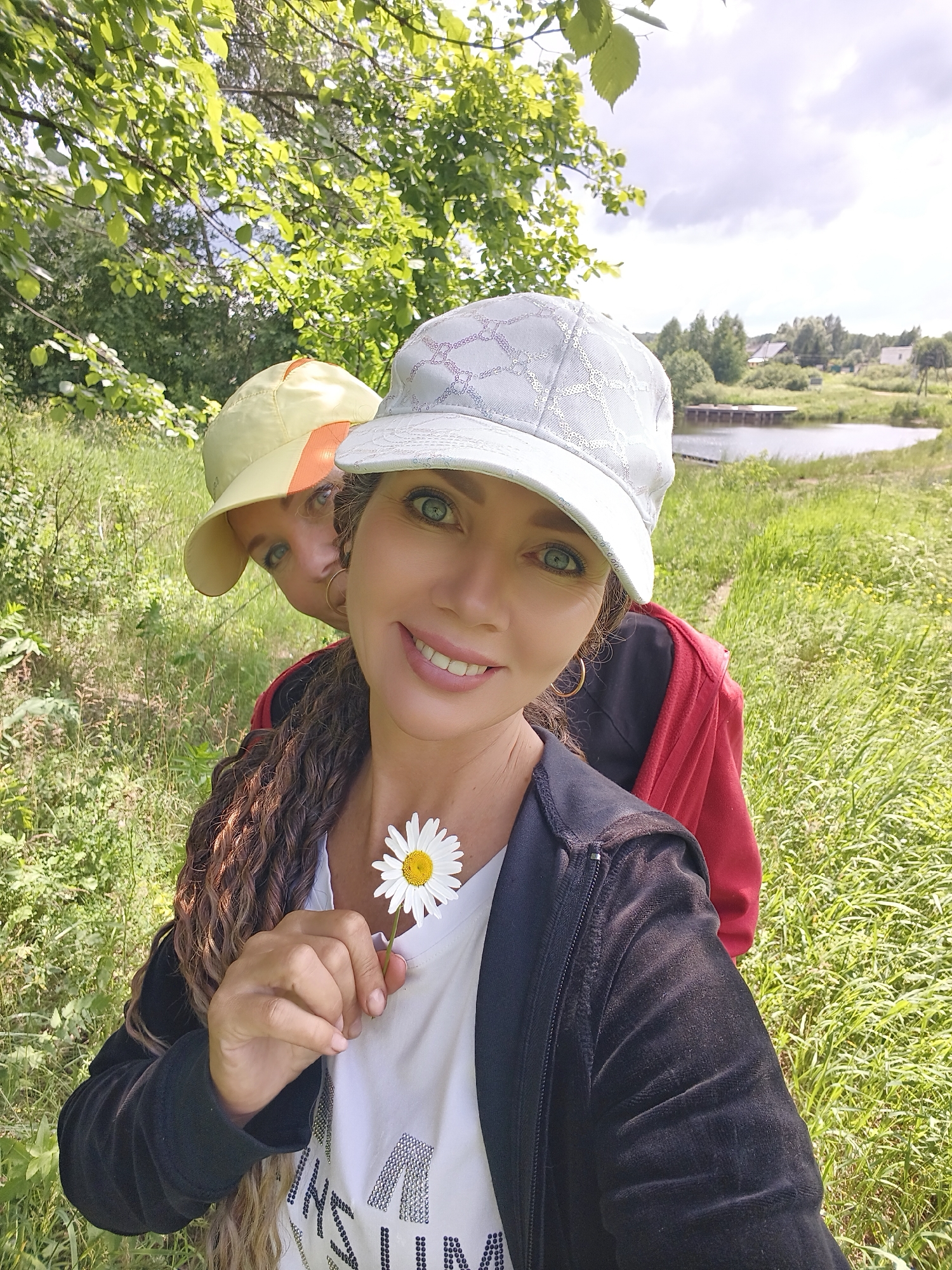 In Zavod Mikhailovskii, Russia, a woman with curly brown hair and striking green eyes, wearing a white t-shirt, a black zip-up jacket, and a light blue baseball cap, holds a daisy up to her chest. Behind her, a child wearing a yellow baseball cap peeks out, their face partially obscured. They are in a grassy outdoor setting with trees and a body of water visible in the background under a cloudy sky. The image is a selfie, captured from a low angle.