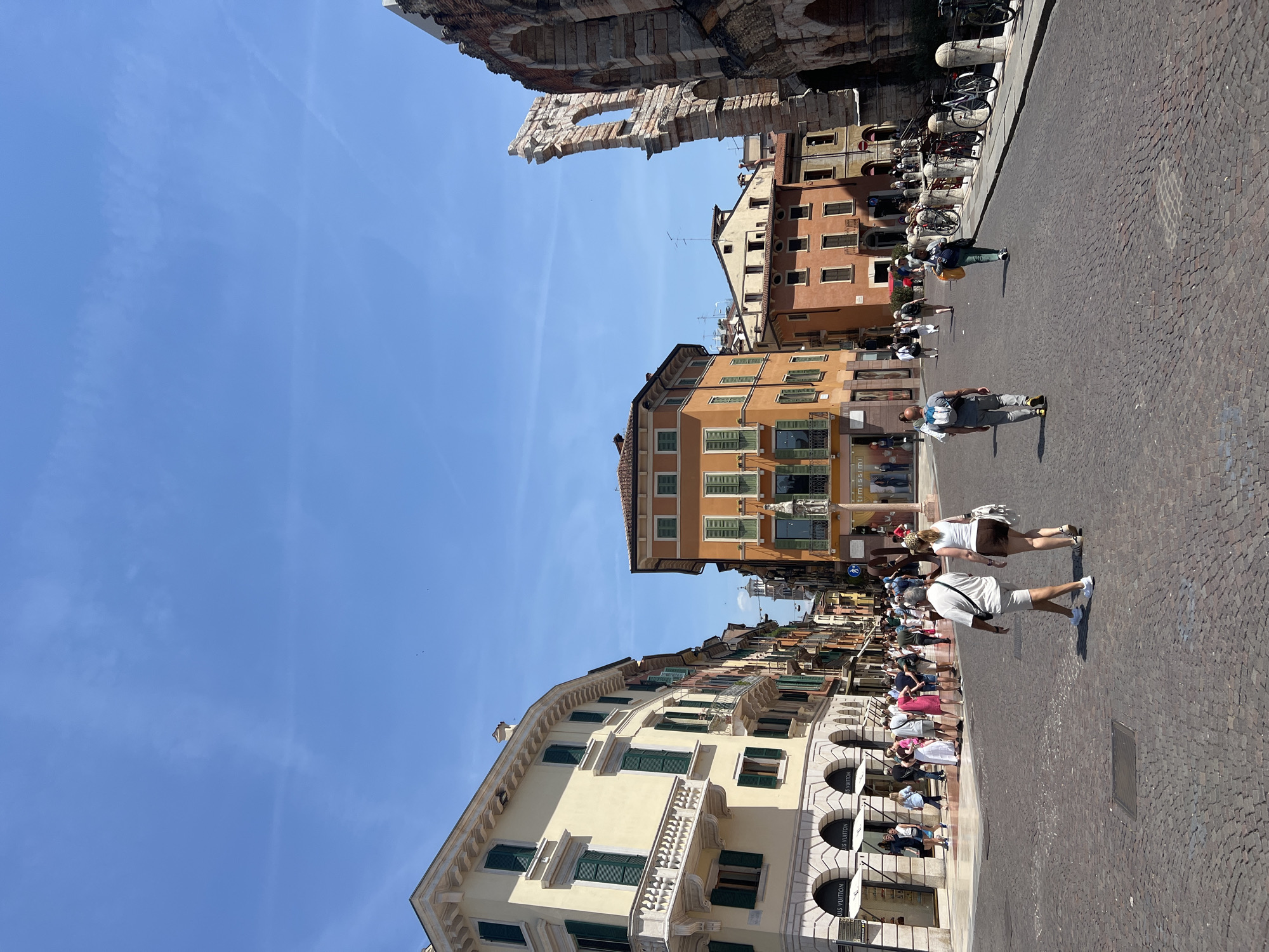 The image shows a view of the Piazza Bra in Verona, Italy, on a sunny day. The foreground features a cobblestone street with a few people walking. On the left side of the street are several historic buildings with light yellow and cream facades, featuring green shutters and balconies. The ground floor of these buildings appears to house shops, including one with "LOUIS VUITTON" signage visible.
Further down the street, on the right side, the edge of the Verona Arena, a well-preserved Roman amphitheater, is visible. The Arena's ancient stone arches and weathered facade are prominent. Behind the Arena, a bright orange building stands out, also with green shutters. Other buildings in shades of orange, red, and yellow line the street, creating a picturesque cityscape. The sky is a clear, bright blue with a few faint wisps of clouds. People are seen strolling, some in groups, contributing to the lively atmosphere of the piazza. Bicycles are parked along a railing near the Arena.