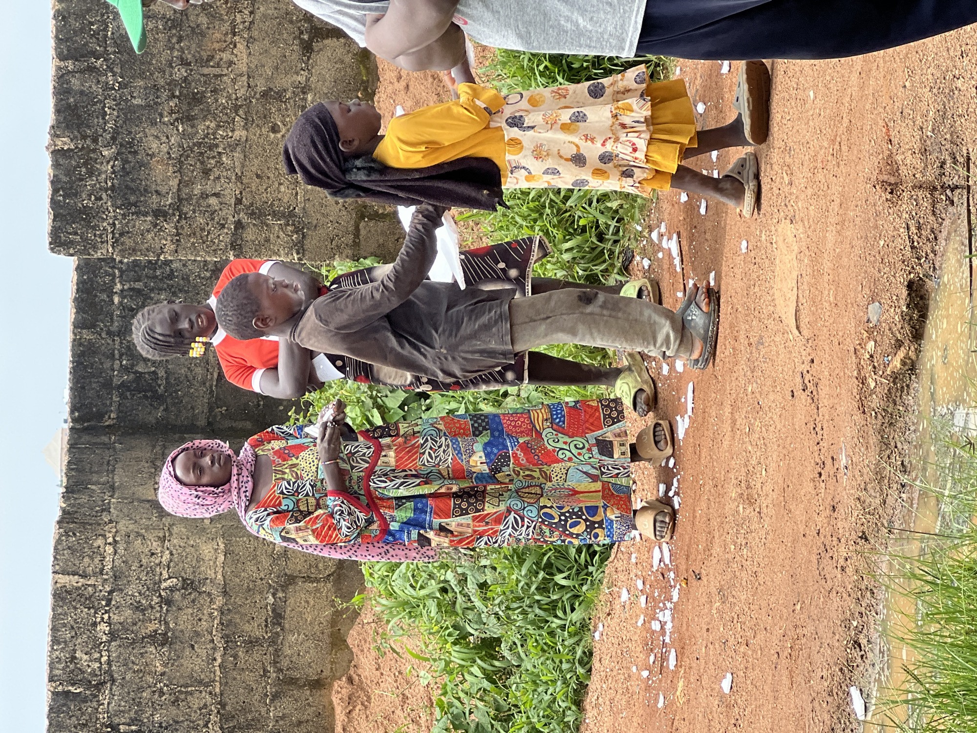In Jos, Nigeria, on a dirt road, a group of four children are gathered near a brick wall. The child on the far left wears a pink headscarf and a colorful, patterned dress. Next to her stands a child in a red shirt with white trim. In the center, a child in a brown shirt and grey pants holds a white object. On the right, a child with a dark headscarf wears a yellow top and a patterned skirt with a ruffled hem. The ground is covered in reddish-brown dirt, with patches of green grass and a small puddle of water. The scene is overcast, with a muted sky.