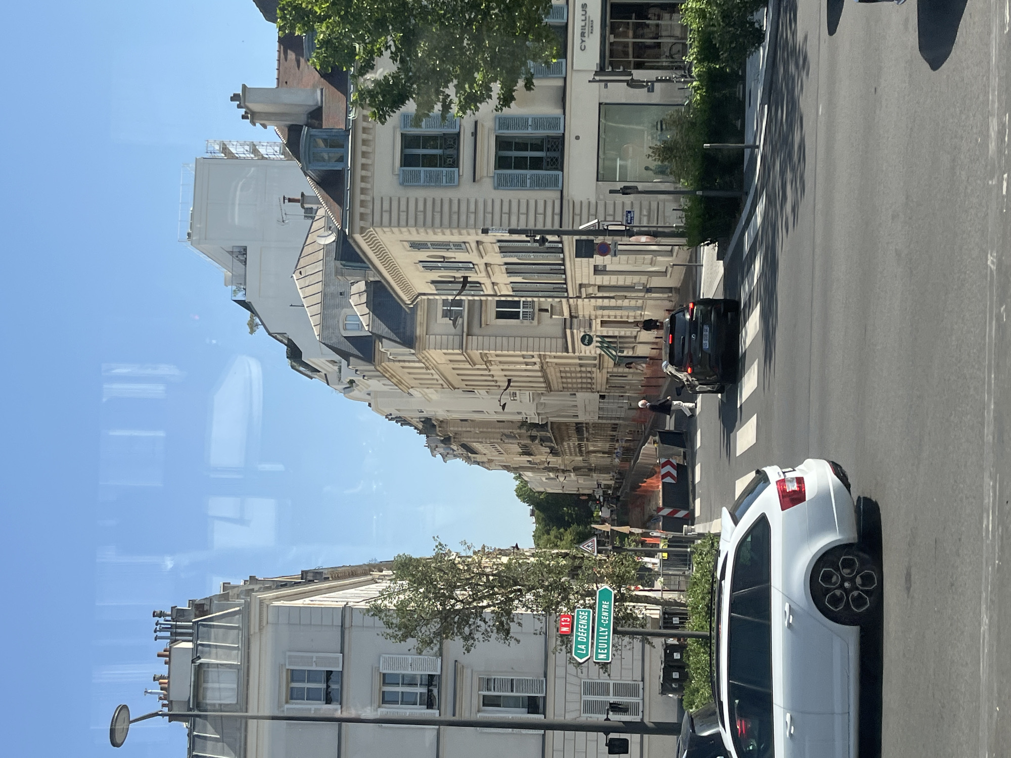 The image depicts a street scene in Neuilly-sur-Seine, France, with a view looking down a tree-lined avenue. On the left, a white apartment building with grey mansard roofs and numerous windows is visible. A street lamp with a circular fixture stands in front of this building. Green street signs indicating "N 13", "LA DÉFENSE", and "NEUILLY-CENTRE" are mounted on a pole.
The right side of the street features a row of cream-colored buildings with light blue shutters, typical of Parisian architecture. These buildings have multiple stories and ornate facades. One building has a shop with a large glass storefront labeled "CYRILLUS".
In the foreground, a white station wagon is driving on the asphalt road. Further down the street, a black SUV is also in motion. There is a crosswalk marked on the road, and temporary orange barriers with reflective tape suggest ongoing construction or roadwork. A few pedestrians are present, walking on the sidewalks or crossing the street. The sky is clear and blue. Reflections are visible on the surface from which the photograph was taken, likely a window.