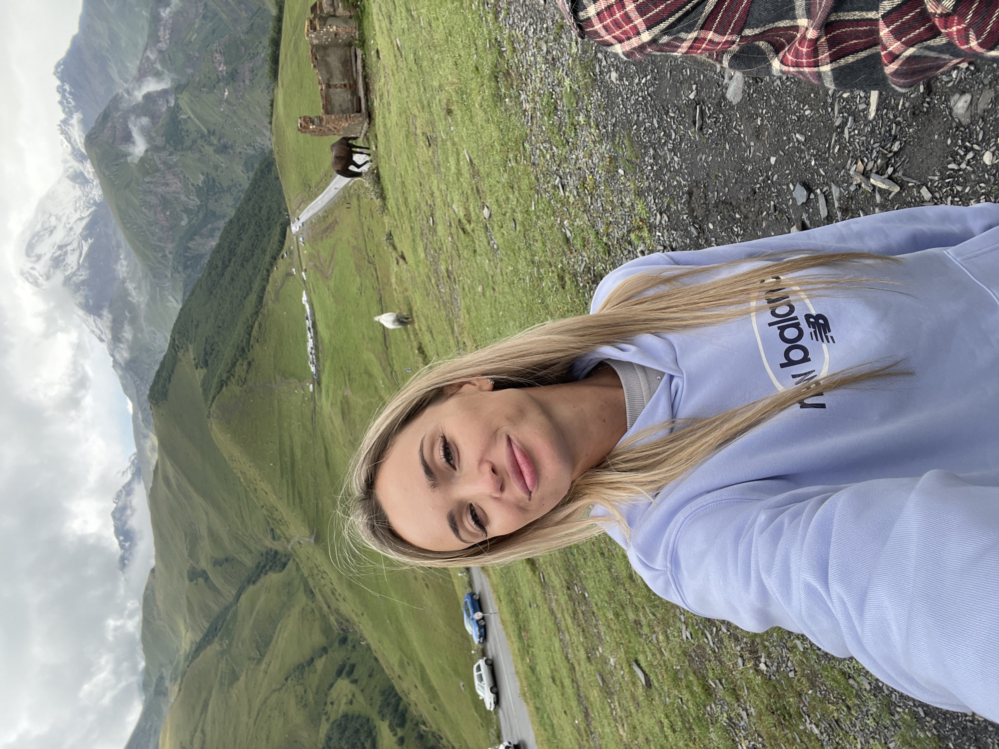 A medium shot captures a young, blonde Caucasian woman taking a selfie in a mountain landscape near Stepantsminda, Georgia. She is positioned in the foreground, looking directly at the camera with a slight smile and her head tilted. She has fair skin, long blonde hair, and is wearing a light blue hooded sweatshirt with a "New Balance" logo. Behind her, the landscape unfolds with rolling green hills, a winding road, several cars parked on the side, and two horses grazing. In the distance, snow-capped mountains loom under a cloudy sky. A partially ruined brick structure is visible on a hill to the right of the frame. The scene suggests a cool, overcast day.
