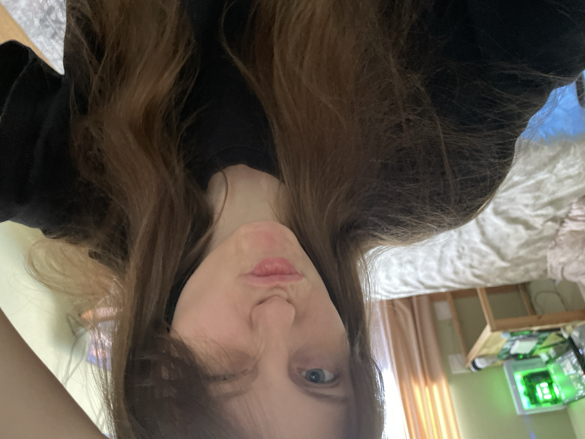 A young woman with long, brown hair is taking a selfie in what appears to be a bedroom. She is wearing a black t-shirt and has her hair falling over her face, with one eye closed and her lips pursed in a pout. In the background, a bed with white and patterned bedding is visible, along with a desk with a computer monitor displaying green graphics. Curtains hang in the background, suggesting a window. The overall lighting is soft, possibly from natural light. The image does not contain any information that would indicate its location in Ufa, Russia.