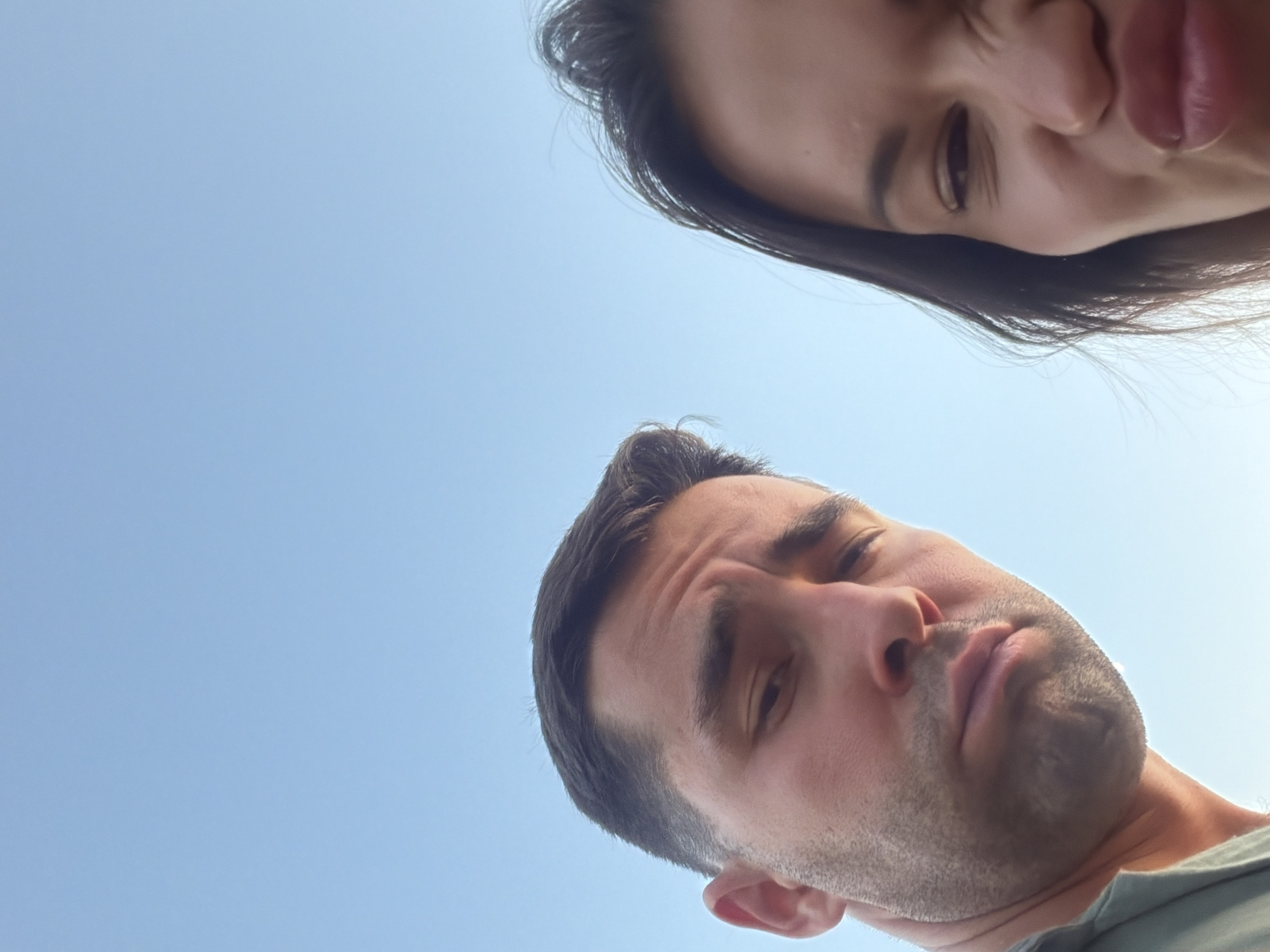 The image is a close-up, low-angle selfie taken outdoors against a clear blue sky. Two individuals are visible: a man on the left and a woman on the right. The man's face is partially obscured, showing his forehead, left eye, nose, and mouth. He has short, dark hair and appears to be making a slightly distressed or bored expression, with his eyebrows furrowed and his mouth downturned. The woman's face is also cropped, revealing her right eye, nose, and mouth. She has long, dark hair and is making a playful or goofy expression, with her eyes squinted and her lips pursed. The lighting suggests it is daytime. The background is uniformly sky, indicating an open outdoor environment, possibly a park or a viewpoint, consistent with the area around Sirmione Castle in Sirmione, Italy, which overlooks Lake Garda.