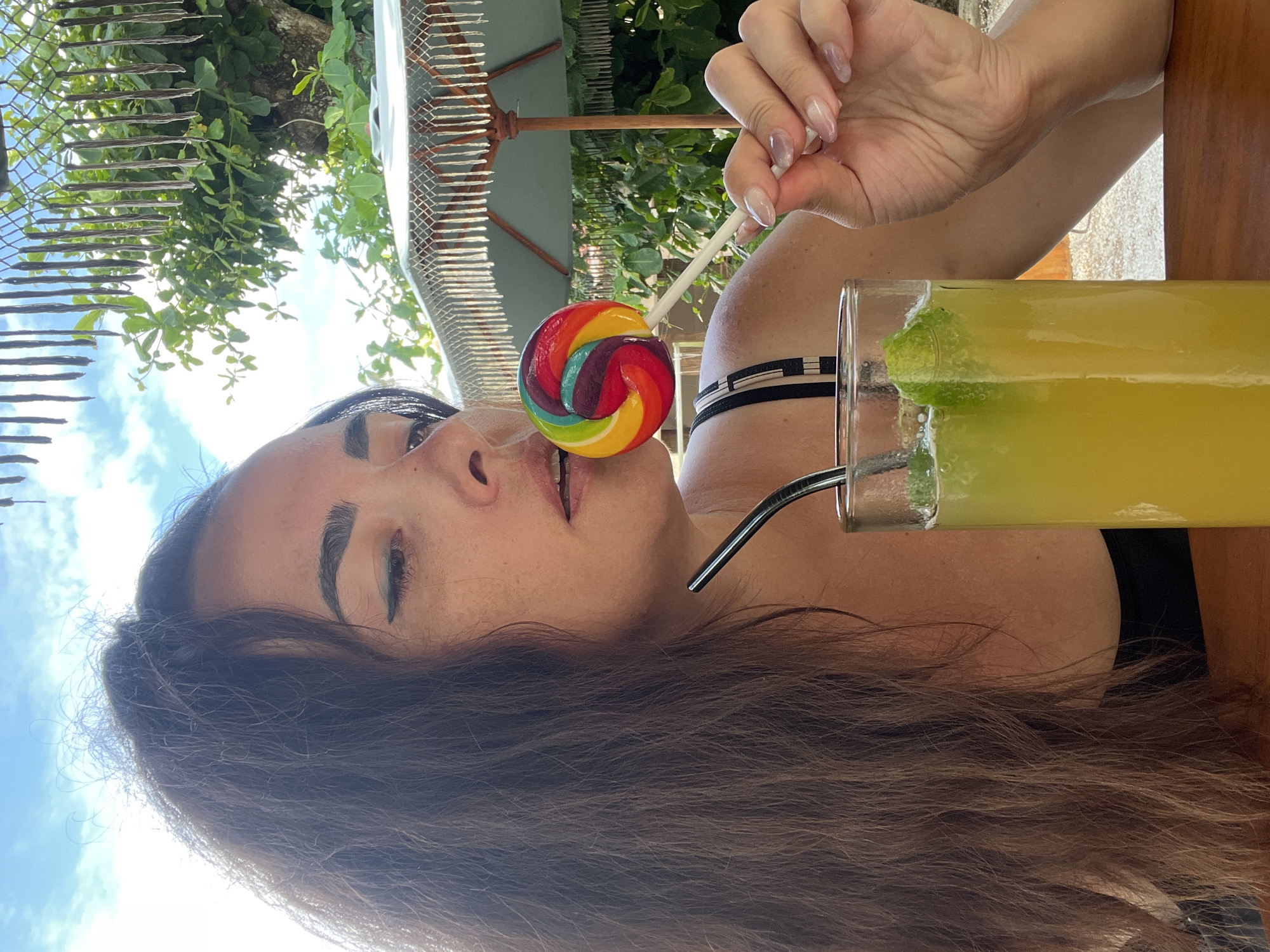 In Kuta Selatan, Indonesia, a woman is captured in a close-up selfie, holding a colorful, swirling lollipop near her mouth. She has long, dark, wavy hair that frames her face, and is wearing a black top. Her eyes are looking away from the camera, and she has noticeable eyeliner. Beside her, a tall glass of a yellow beverage, possibly a lime-based drink with mint, is visible, with a metal straw inserted. In the background, a lush green tree provides a natural backdrop, with a fringed umbrella and a clear blue sky above. The setting suggests an outdoor cafe or restaurant.