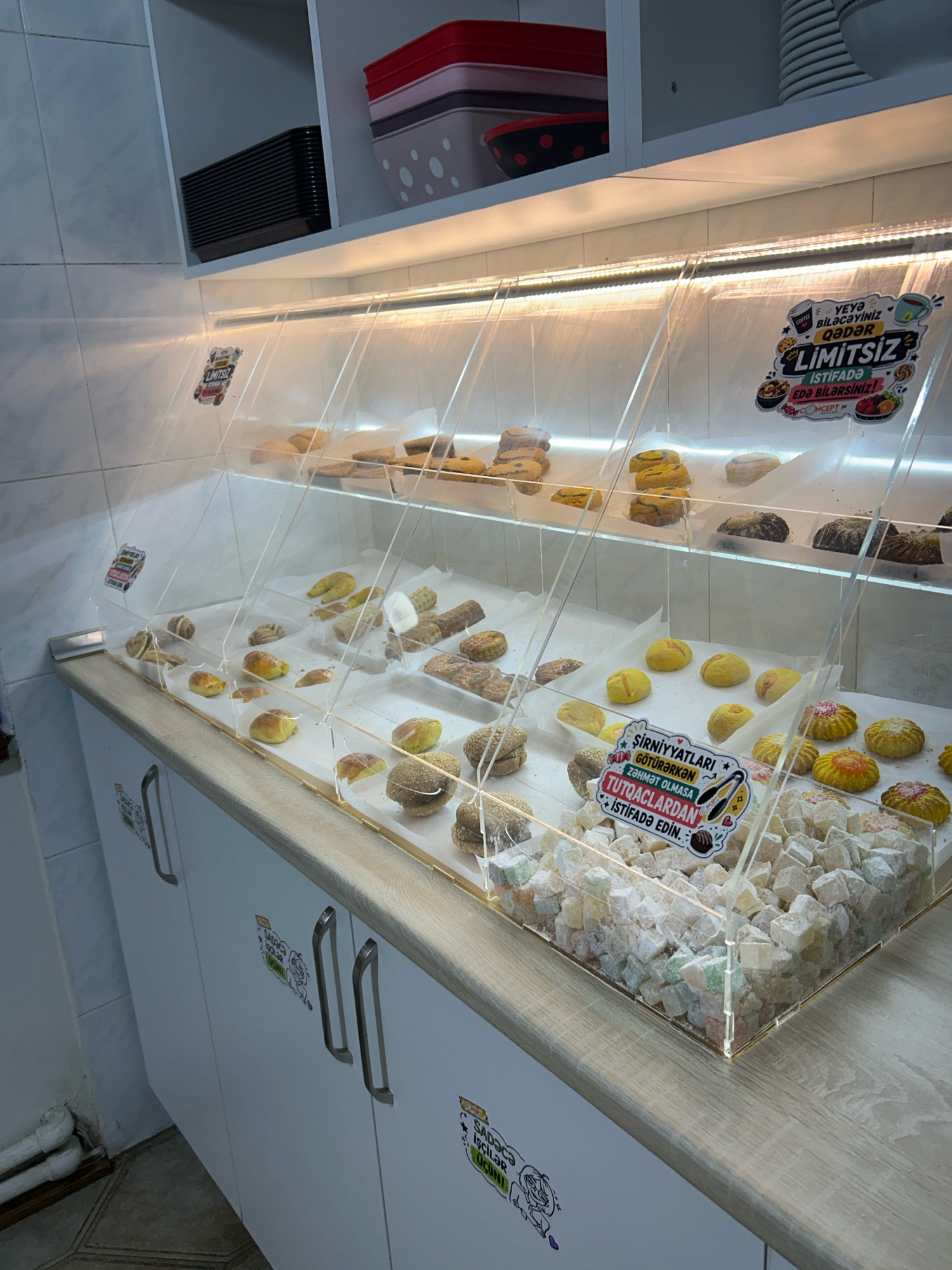 This is a photograph of a confectionery display case filled with various types of pastries and Turkish delight. The display case is made of clear acrylic and is illuminated by an LED strip light. The pastries on display include cookies, small baked goods resembling croissants, and round, textured items. In the foreground, a large quantity of Turkish delight, cut into cubes and dusted with powdered sugar, is presented in a rectangular acrylic tray.
Two signs are visible within the display case. One, at the top, has an image of coffee and cookies and reads "YEYƏ BİLƏCƏYİNİZ QƏDƏR LIMITSIZ İSTİFADƏ EDƏ BİLƏRSİNİZ!" which translates to "You can use as much as you can eat, unlimited!". The other sign, in the middle, features cartoonish illustrations and text that appears to be in Azerbaijani, with phrases like "ŞİRNİYYATLARI GÖTÜRƏRKƏN ZƏHMƏT OLARSA TUTQACLARDAN İSTİFADƏ EDİN." translating to "When taking sweets, please use tongs."
On the cabinetry below the display, further signage is visible, including one that says "SADƏCƏ İŞÇİLƏR ÜÇÜN" which translates to "For staff only".
The environment appears to be an indoor setting, possibly a bakery or a confectionery shop. The background includes white tiled walls and shelving units holding stacked containers. The overall impression is of a well-stocked and organized retail space. The specific location in the city of Baku, Azerbaijan cannot be determined from the visual information alone.