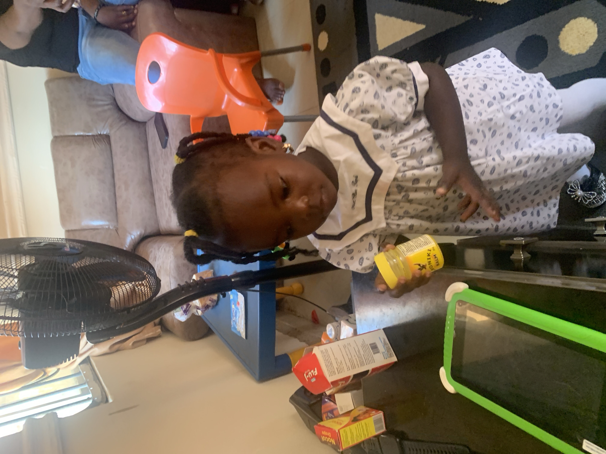 A young Black girl with braided hair stands in a room, holding a yellow jar. She is wearing a white dress with blue trim and a pattern of blue shapes. In front of her is a dark surface with a bright green tablet in a protective case. Behind her, a black oscillating fan stands over a blue table, on which are several boxes, including one red and white box labeled "Flui". Further back, a light brown couch is visible, as is an orange plastic chair. The room appears to be a domestic indoor setting, likely a living room.