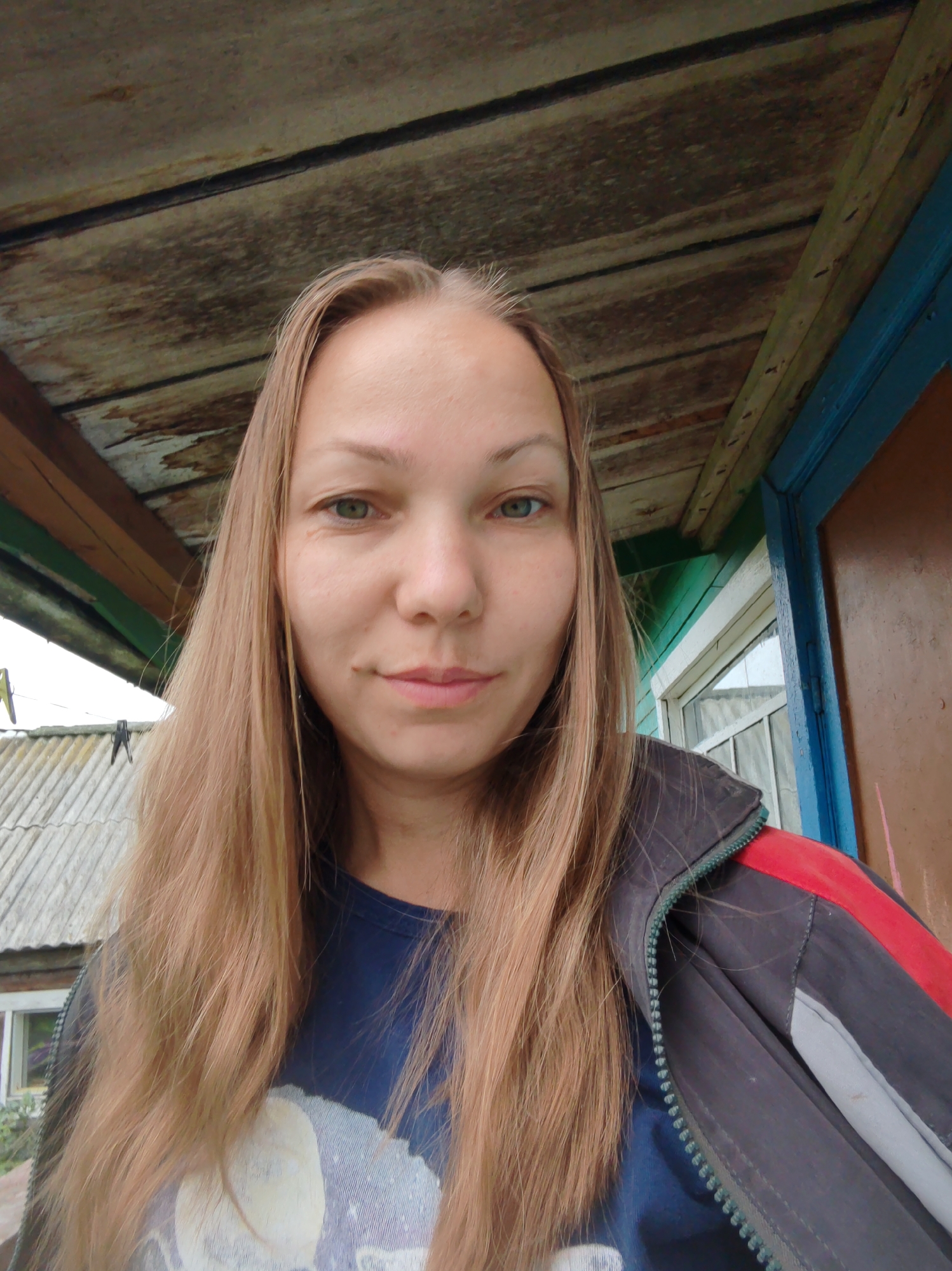 A medium shot captures a fair-skinned woman with long, light brown hair. She is positioned outdoors, with a rustic, wooden ceiling overhead. To her right, a brown door with a blue frame is visible, and a green-painted wall with a window is present. In the background to the left, a house with a gray, corrugated roof and a white-framed window can be seen. The woman is wearing a dark blue t-shirt and a dark jacket with red and gray accents. The scene appears to be in the city of Chuchloma, Russia, suggested by the rural architectural style.