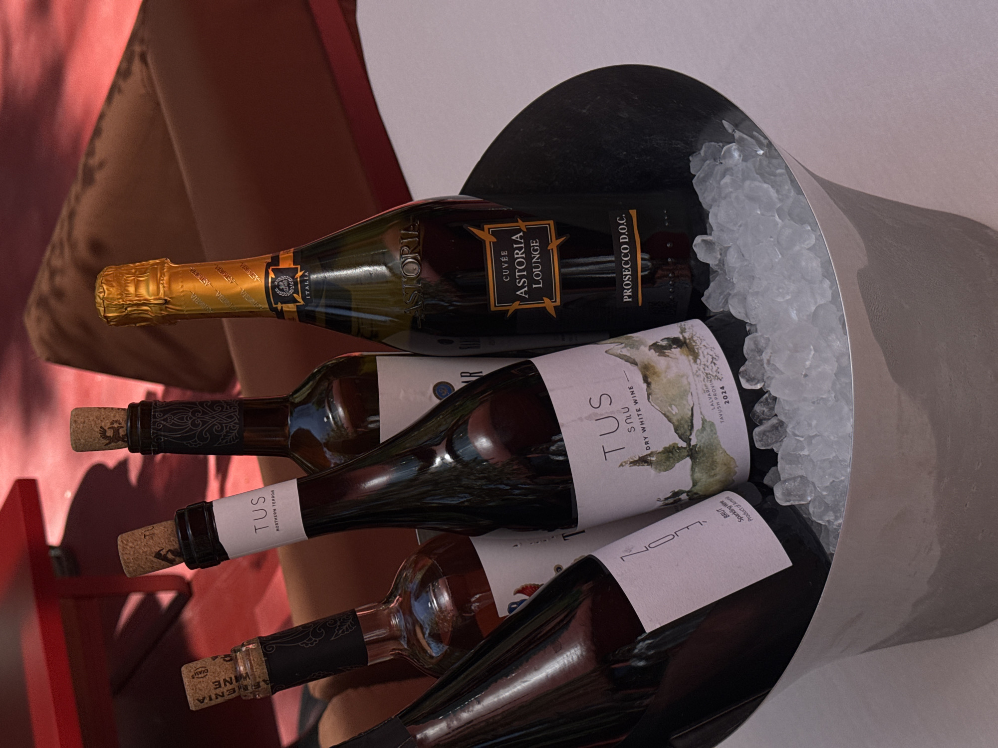 The image shows a metal ice bucket containing four bottles of wine, likely in Yerevan, Armenia, as indicated by one of the wine labels. The top bottle is a gold-foiled Astoria Lounge Prosecco D.O.C. Next is a white wine labeled "TUS", with the text "DRY WHITE WINE" visible. Underneath is another bottle of "TUS" wine. The bottom bottle is partially visible with the label "NOM" and "Sparkling Wine, Product of Armenia" and "AFRENIA WINE" near the cork. Ice fills the bucket around the bottles. The bucket is situated on a surface adjacent to a red chair and a cushion.