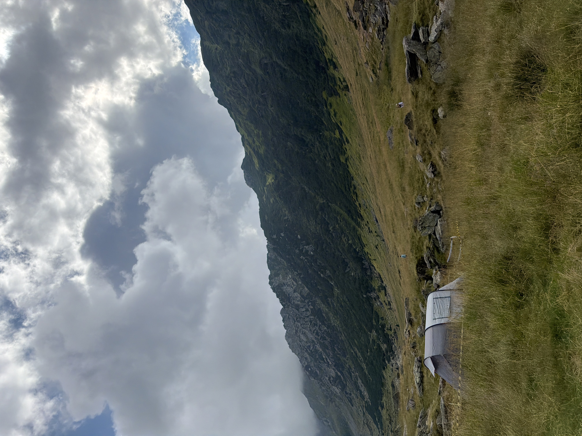 The image depicts a mountainous landscape near Cârțișoara, Romania. A gray and white tent is pitched in a grassy field dotted with rocks. In the background, a steep, green mountainside rises towards a cloudy sky. Two individuals, appearing as small figures in the distance, are walking in the field. The overall scene suggests a remote, natural setting likely used for camping or hiking.