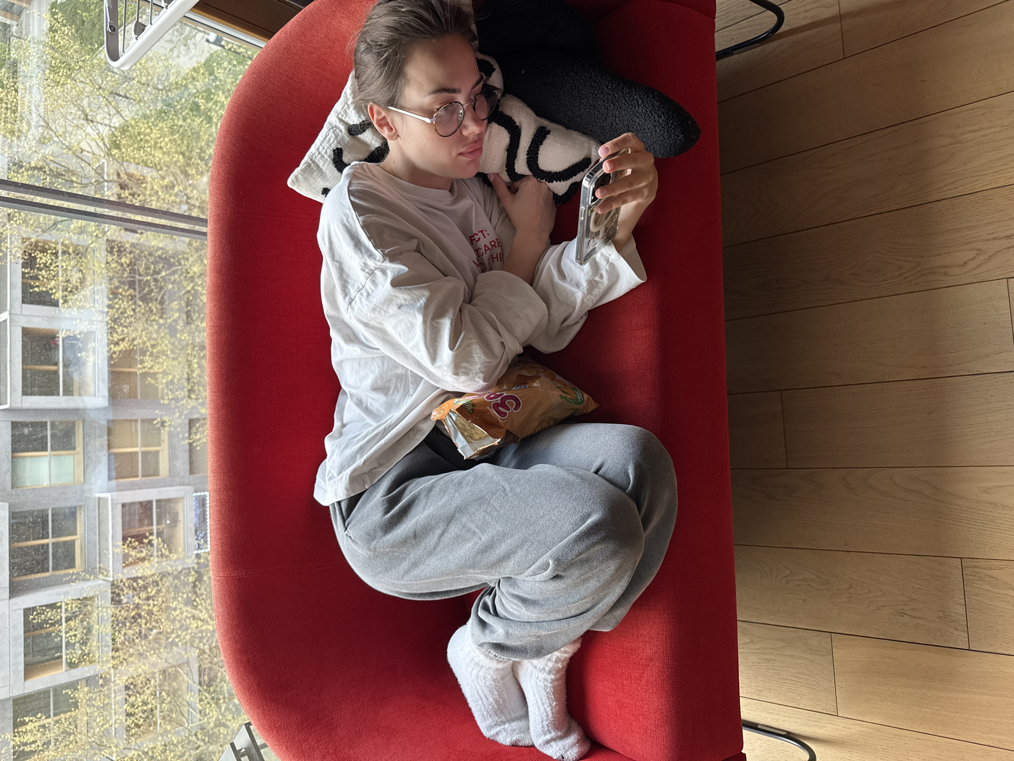 A young person is shown from an overhead perspective, lounging on a bright red upholstered chaise lounge or large armchair. The individual, who appears to be female, has short brown hair pulled back, and wears round eyeglasses. She is dressed casually in a loose-fitting white long-sleeved t-shirt with partial text "CT: CARE MY H" visible, gray sweatpants, and fluffy white socks. She is lying on her left side, resting her head on a white pillow with black abstract patterns, and another black fluffy pillow is behind her.
Her attention is fully captured by a smartphone, which she holds in both hands, positioned directly in front of her. A bag of snacks, possibly "3D's" chips, rests on her lap. The bright natural light streaming in suggests it is daytime. To the left, a large window reveals an outdoor urban scene featuring a multi-story building with many windows and trees displaying fresh, light green foliage, indicating either spring or early summer. To the right, a polished wooden floor is visible. The overall scene depicts a moment of relaxed leisure indoors, likely within an apartment in Warsaw, Poland.