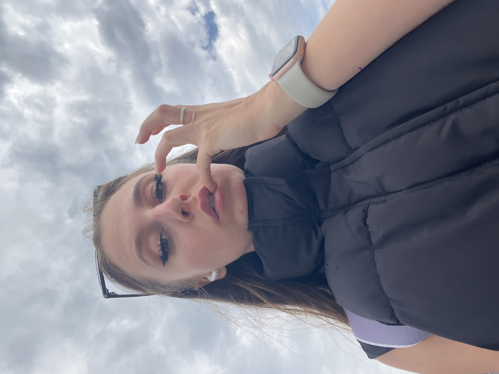 A young person, likely female, is featured in a low-angle shot against a cloudy, overcast sky in Budva, Montenegro. She is looking directly at the camera with a neutral expression, her lips slightly parted. Her left hand is raised, with the index finger and thumb gently touching her face near her mouth or nose.
She wears a dark, possibly black, puffy vest or jacket, with a light purple garment visible underneath. Her accessories include a silver ring on the ring finger of her left hand and a smartwatch with a white band and a rose gold/pink casing on her right wrist. A white wireless earbud is visible in her right ear, and dark-framed glasses rest on top of her head in her hair. Her eyes are enhanced with visible eyeliner and long eyelashes.
The background consists solely of dense, gray clouds, indicating daytime during overcast weather. There are no clear location cues other than the sky itself. No legible text is visible on any items or in the background.